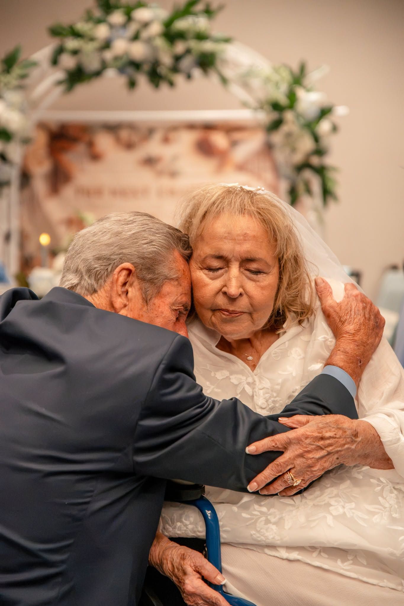 Elderly couple sharing a tender embrace at their wedding ceremony, bride in white veil, groom in blue suit.