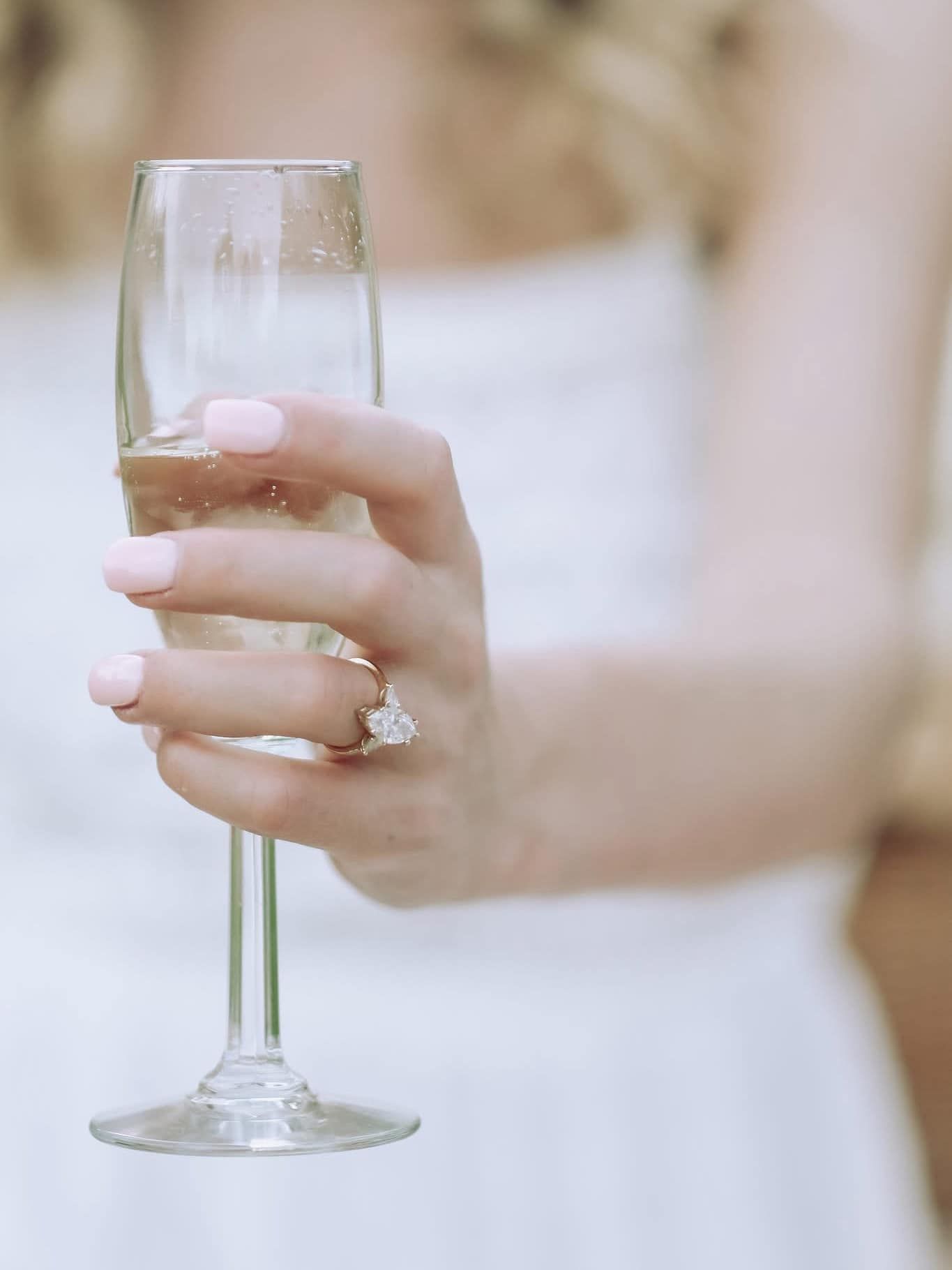A bride with pink nails and diamond ring holds a champagne flute in a white dress.