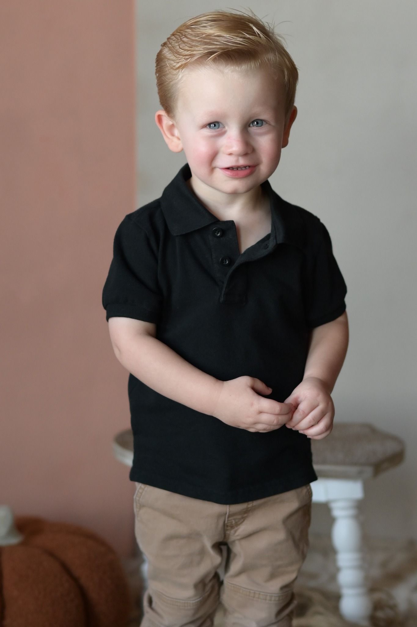 An adorable toddler boy with blue eyes smiles in a black polo shirt and khaki pants.