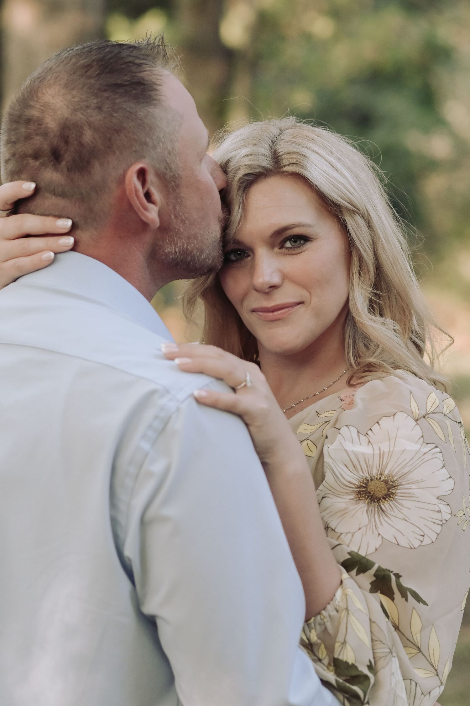Woman in floral dress smiling softly as man kisses her cheek during outdoor engagement photo session.