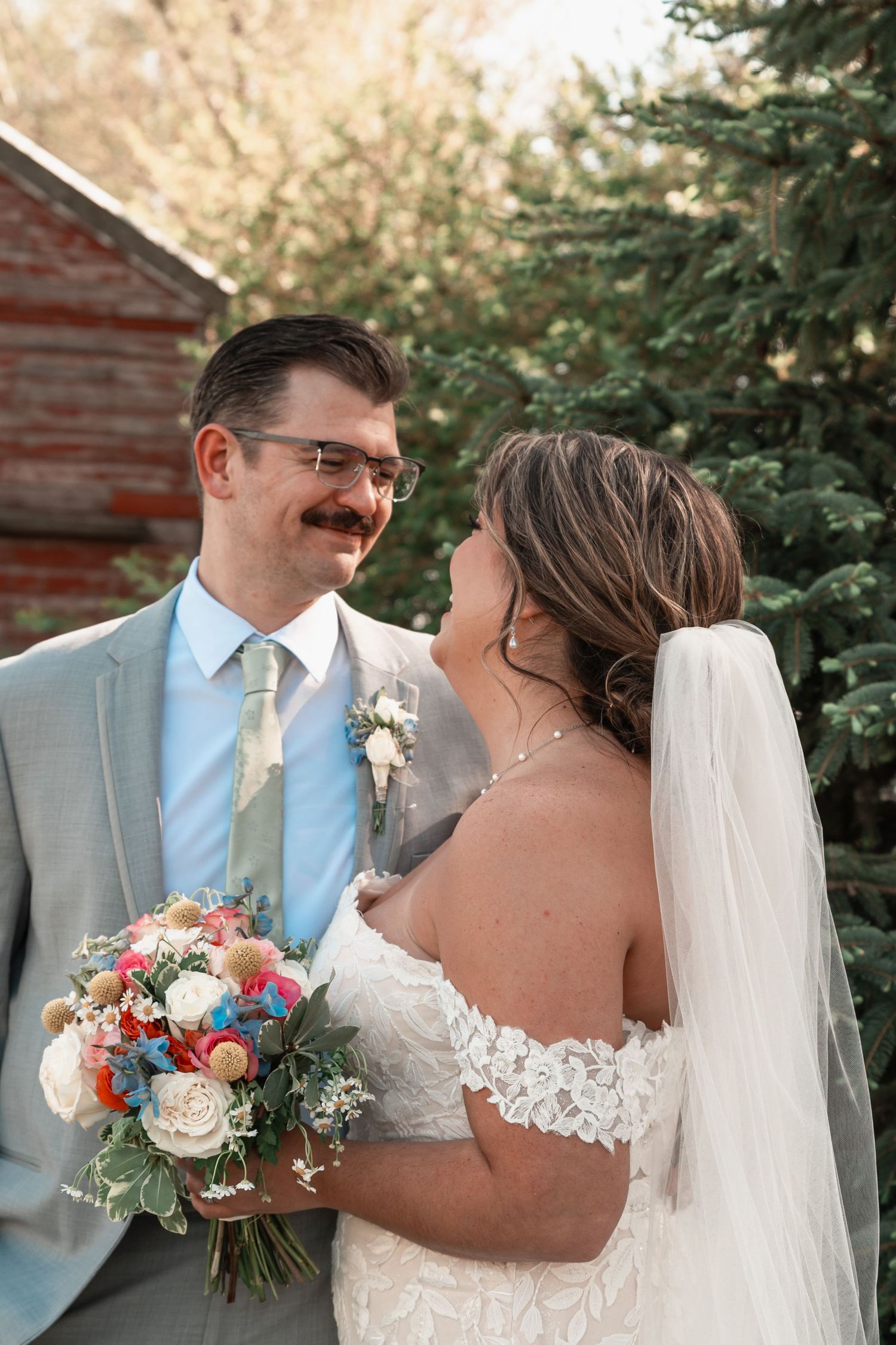 Bride and groom share a tender moment outdoors, she holds a colorful bouquet, he wears a gray suit with sage tie.