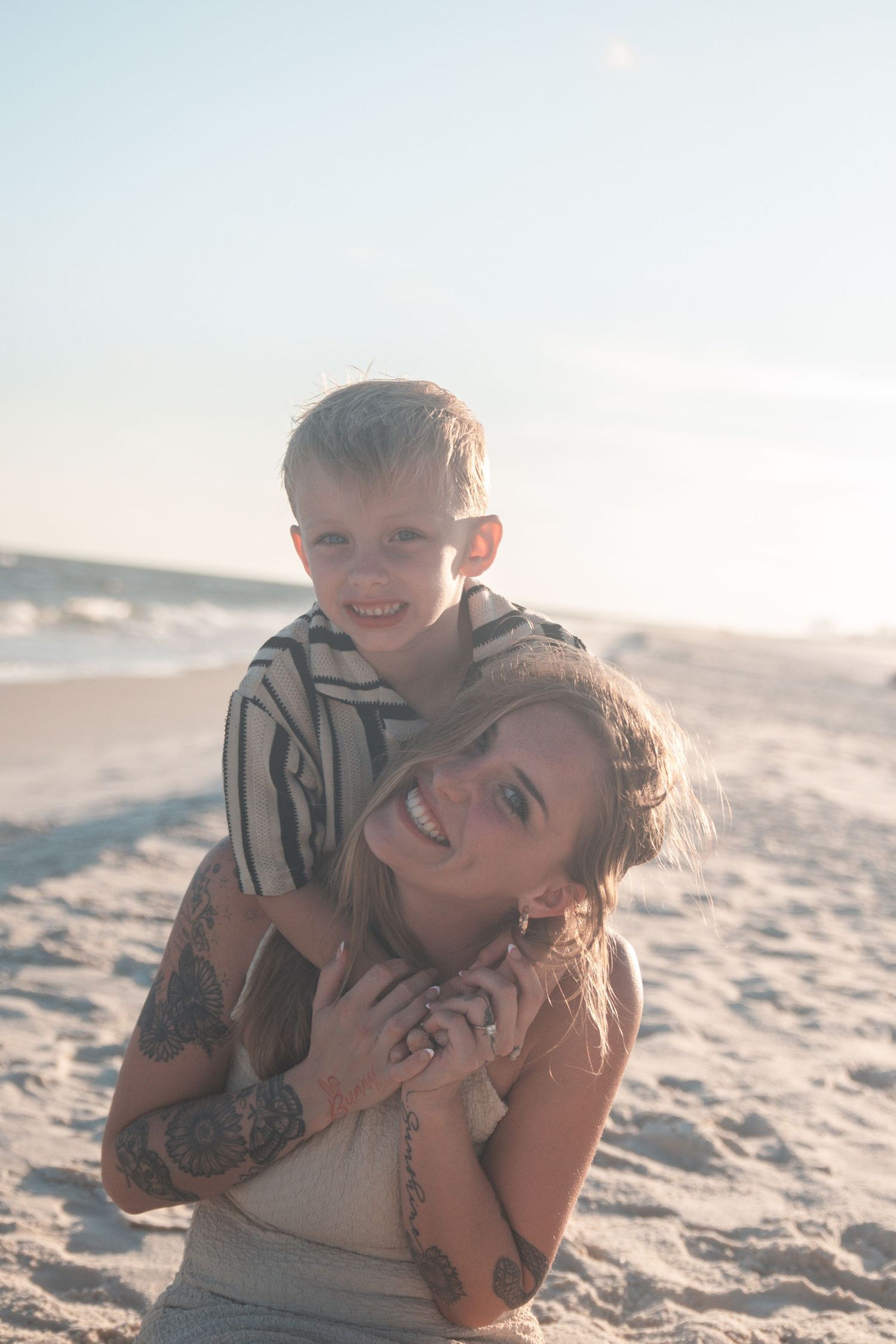 A tattooed woman smiles on a sunny beach while a young blonde boy hugs her from behind.