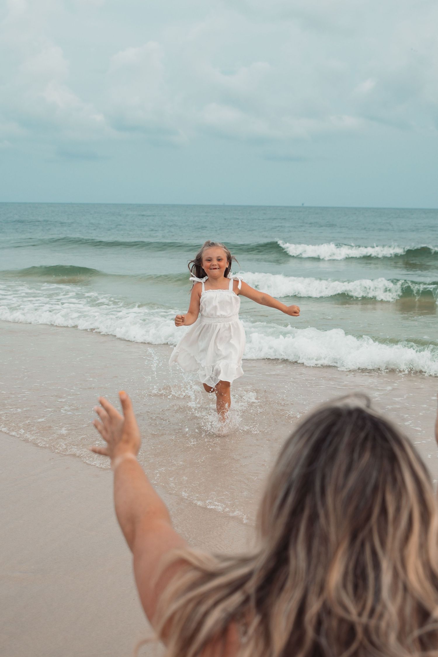 A joyful little girl in a white dress runs along the beach toward outstretched arms, with ocean waves behind her.
