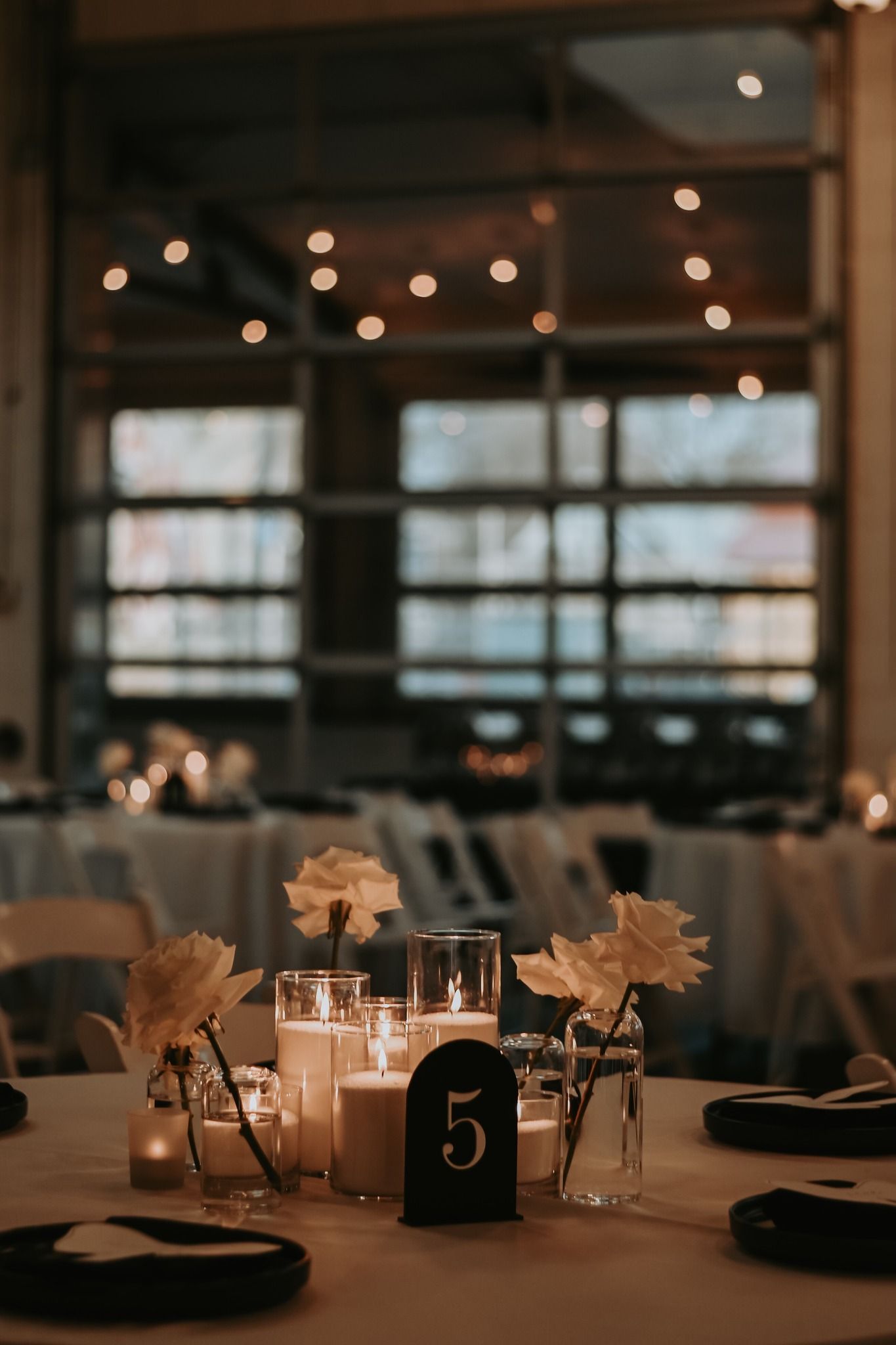 Soft-lit wedding table centerpiece featuring white roses, glowing candles, and elegant black number 5 card.