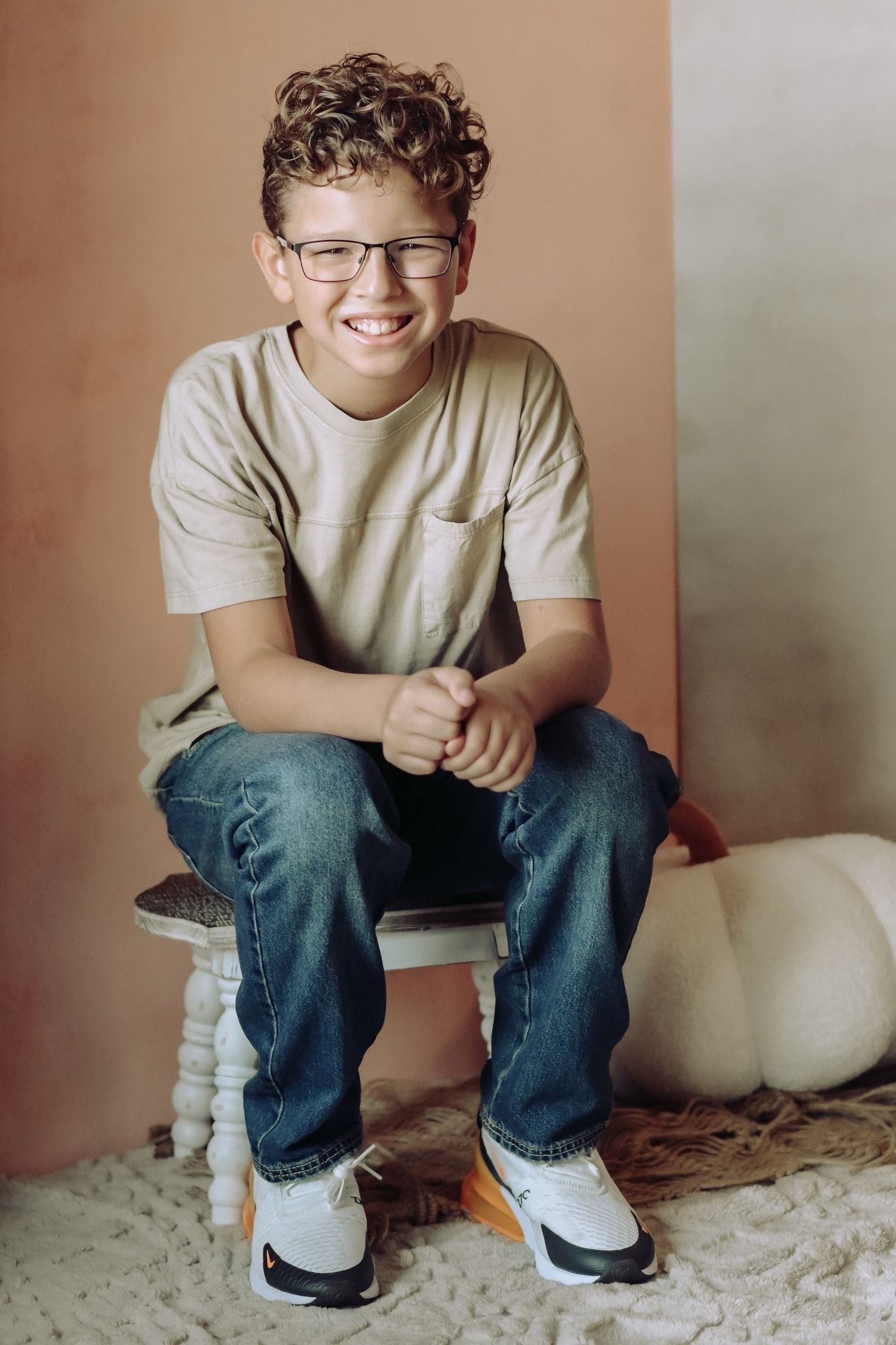 A smiling curly-haired boy with glasses sits on a stool in jeans and a beige t-shirt.