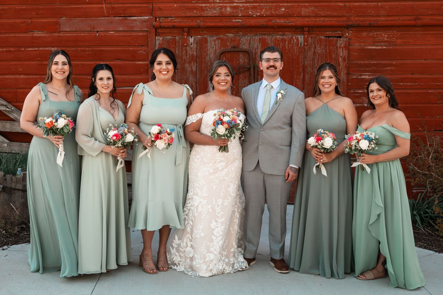 Wedding party posing in front of red barn with bridesmaids in sage green dresses and groom in gray suit.
