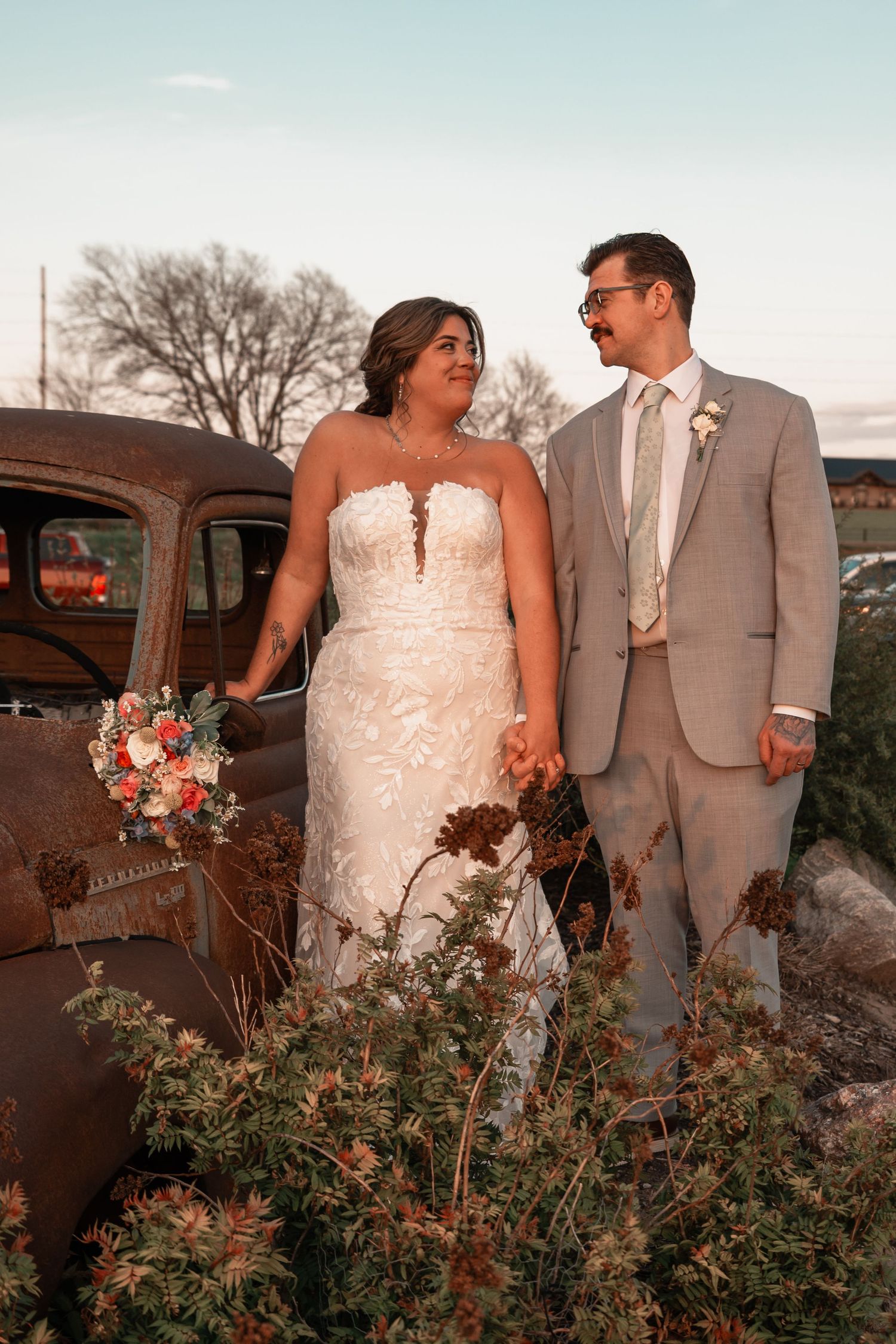 Bride and groom pose beside rustic vintage truck at sunset with colorful bridal bouquet.