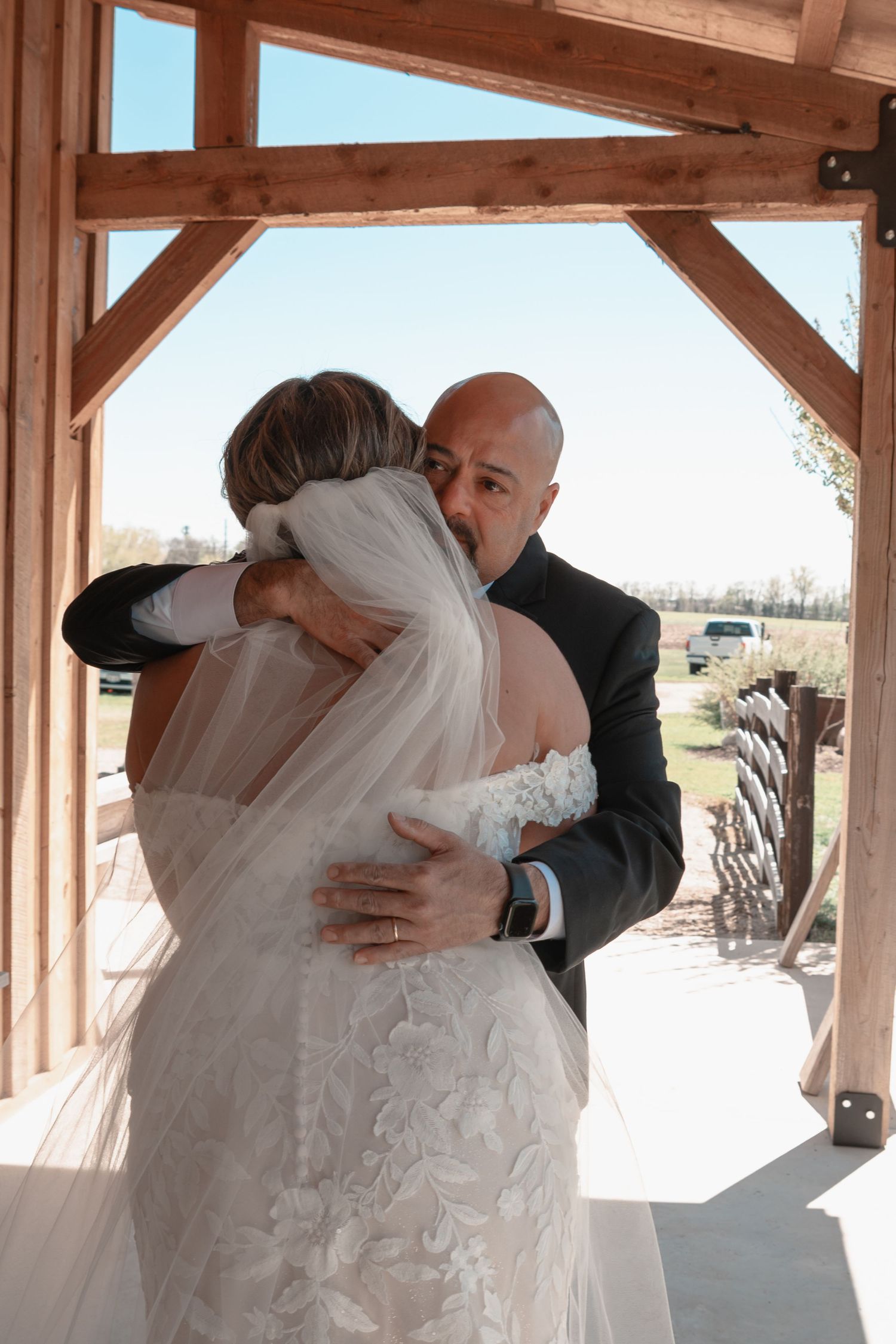 Emotional first look embrace between bride in lace gown and bald man under rustic wooden pergola.