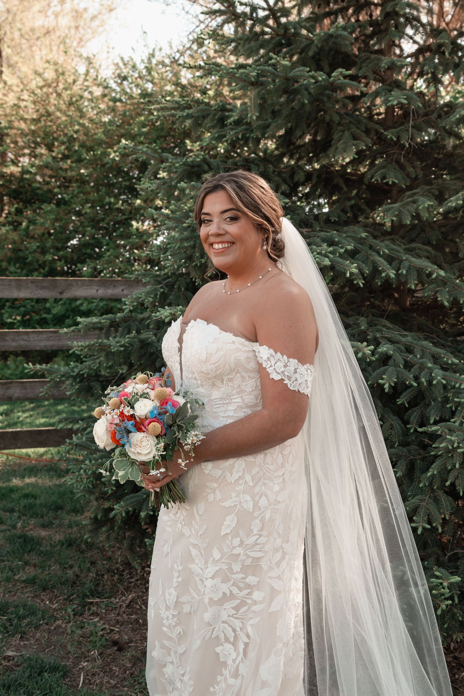 Radiant bride in off-shoulder lace gown holding colorful bouquet near pine trees.