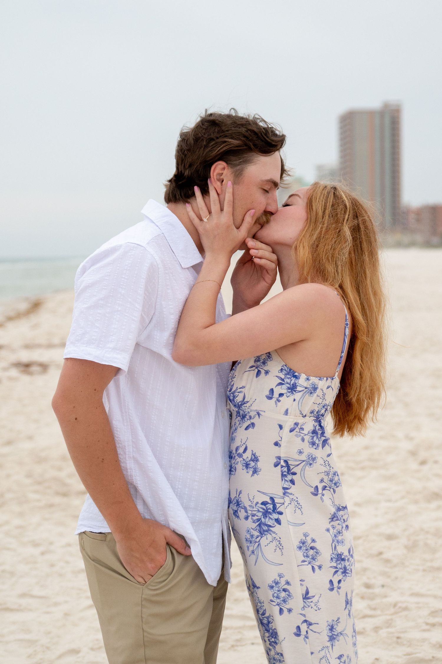 A couple kisses tenderly on a sandy beach with a city skyline visible in the distance.