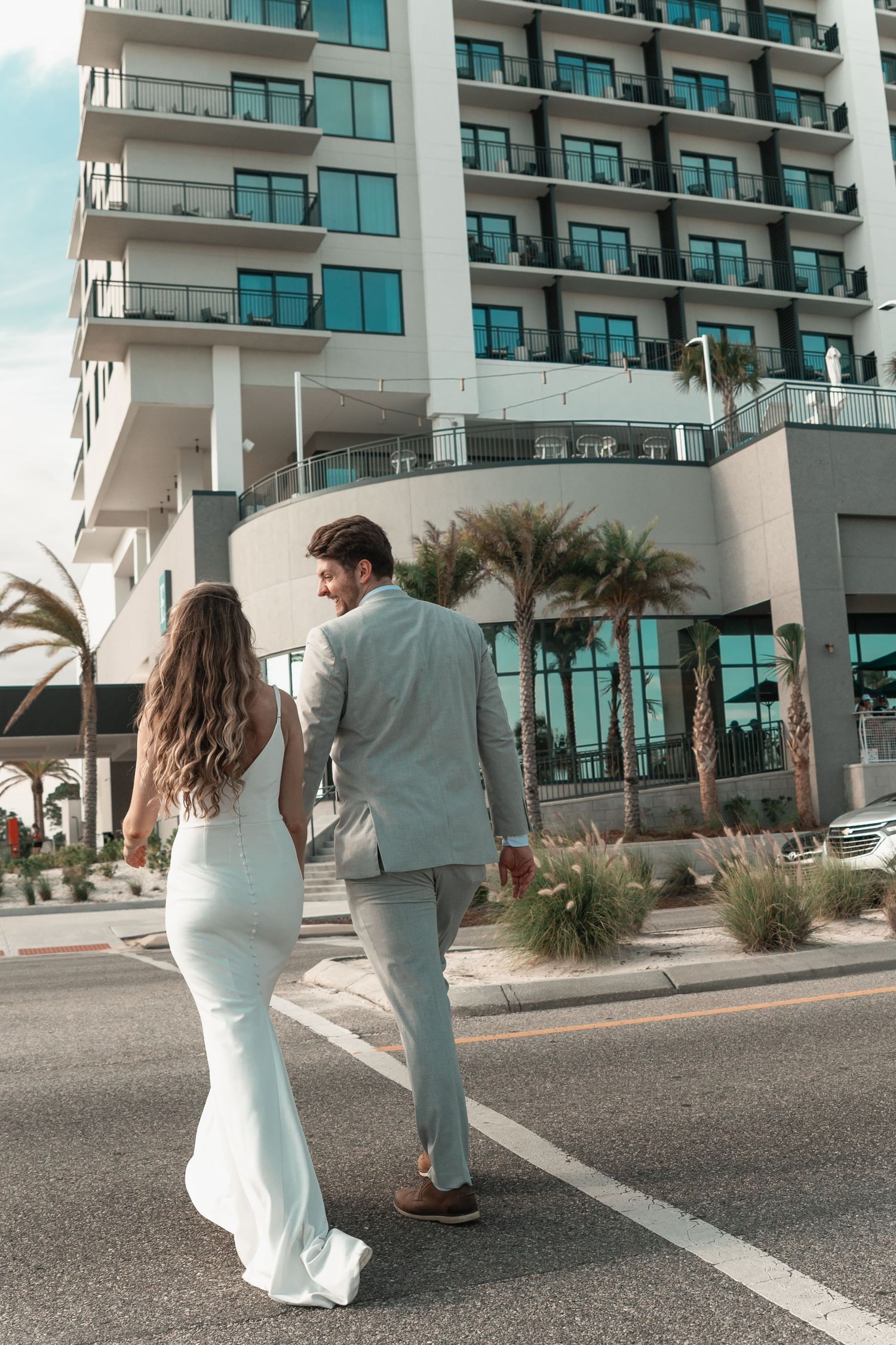 A bride and groom in white attire walk hand in hand past a modern high-rise building lined with palm trees.