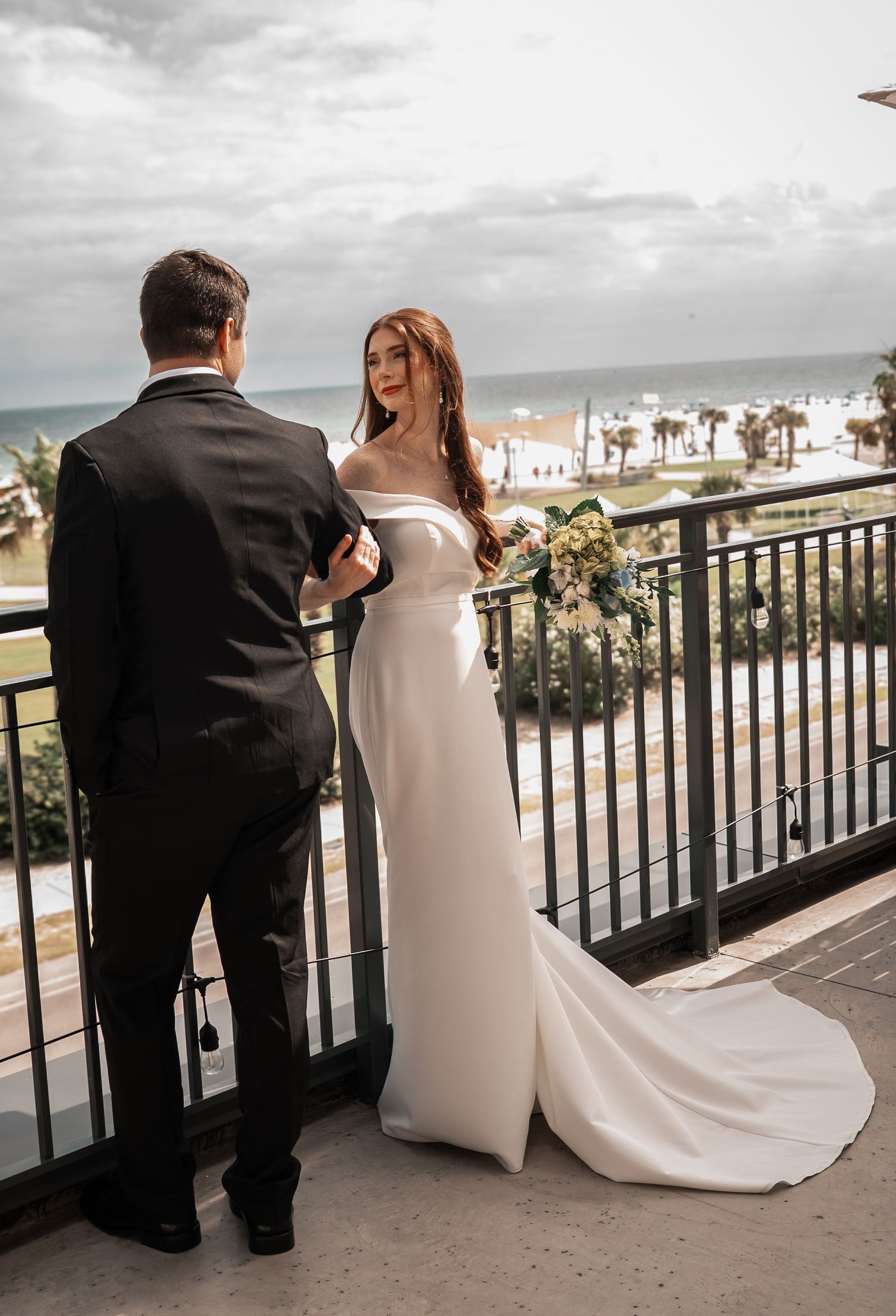 Elegant bride in off-shoulder white gown with train standing on balcony overlooking ocean.