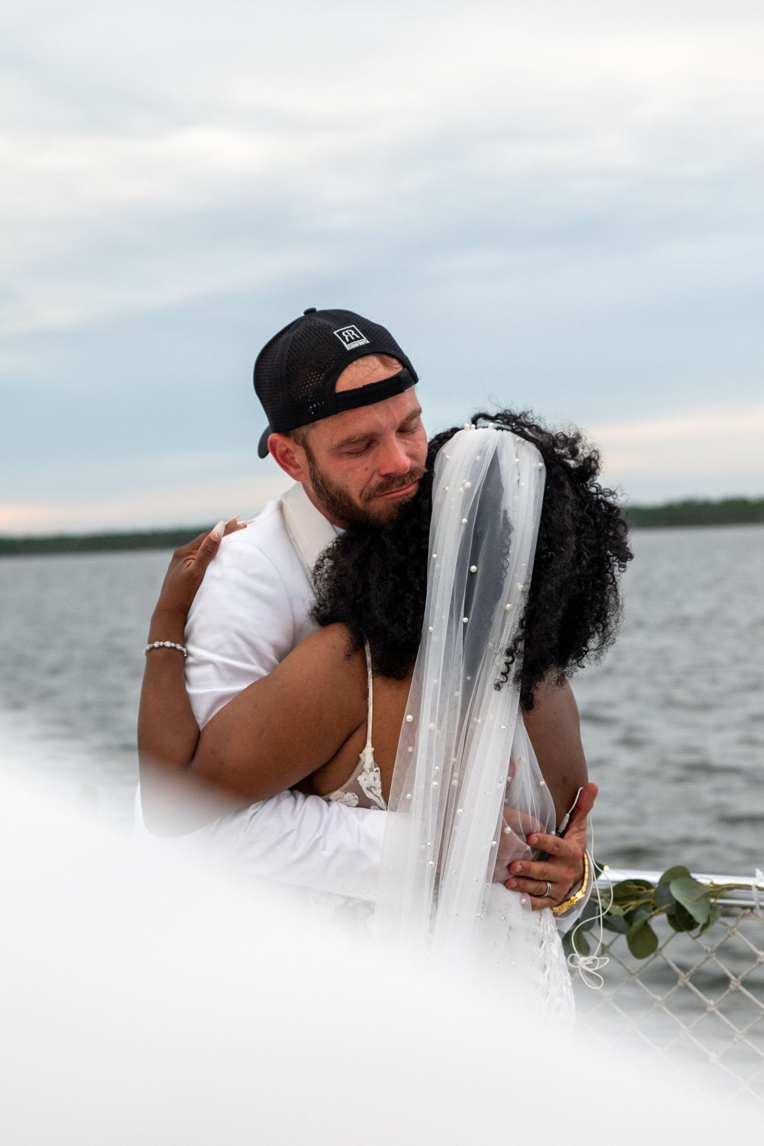 A couple shares a romantic embrace on a boat, the bride wearing a veil and groom in a white shirt and black cap.