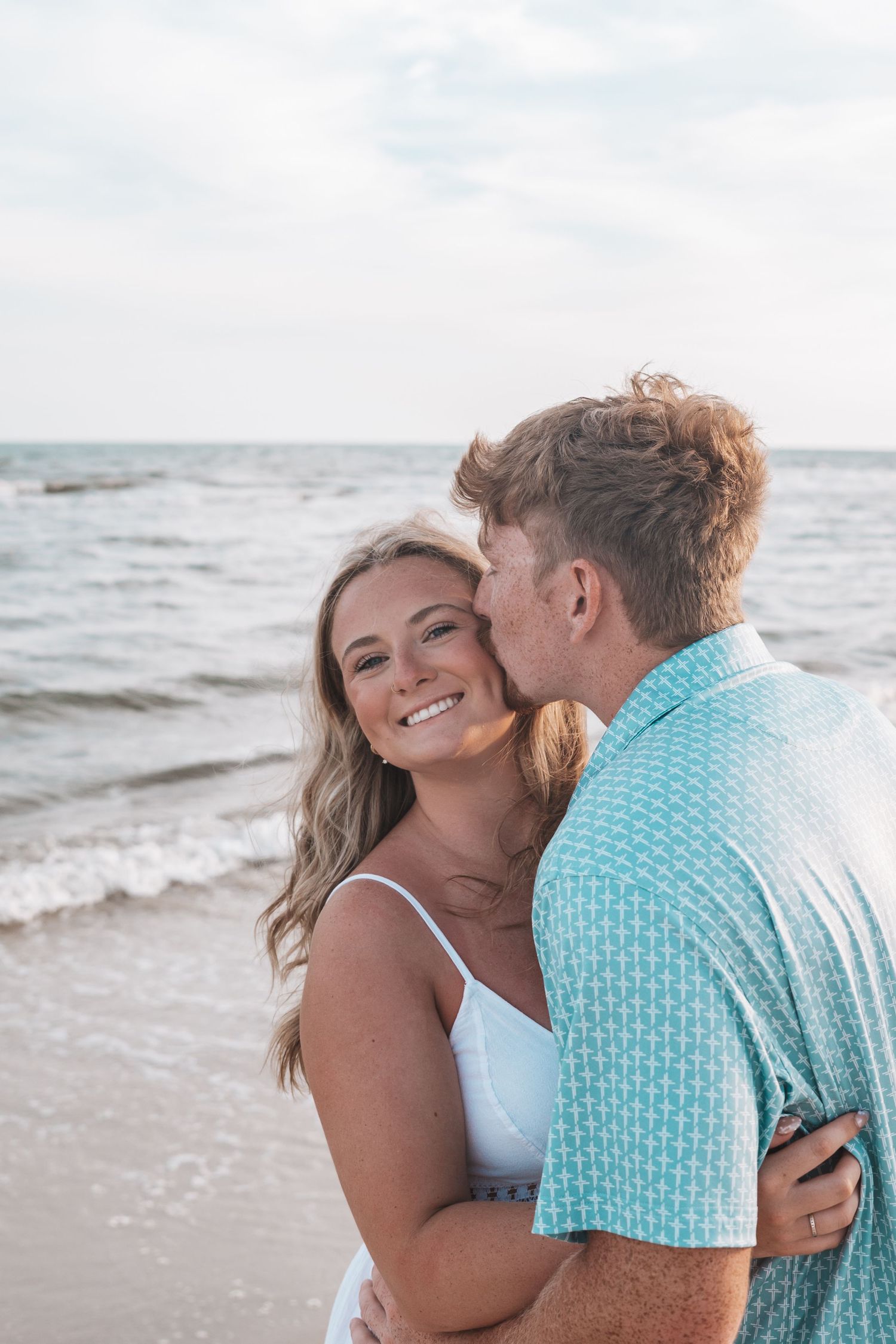 A couple embraces on a beach, woman smiling at camera as man kisses her cheek, ocean waves in background.