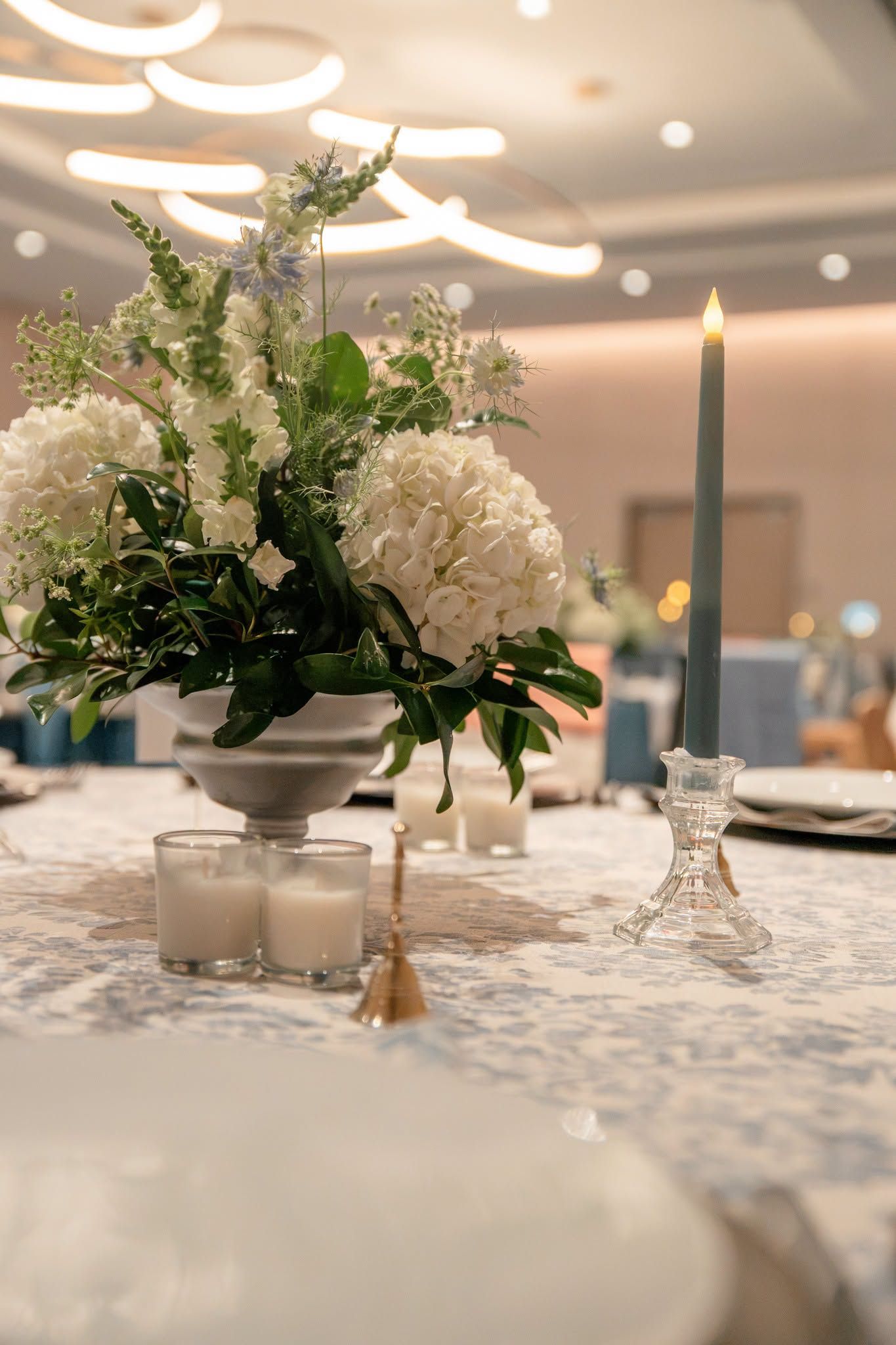 Elegant wedding table with white hydrangea centerpiece, taper candle, and votive candles on a blue floral tablecloth.