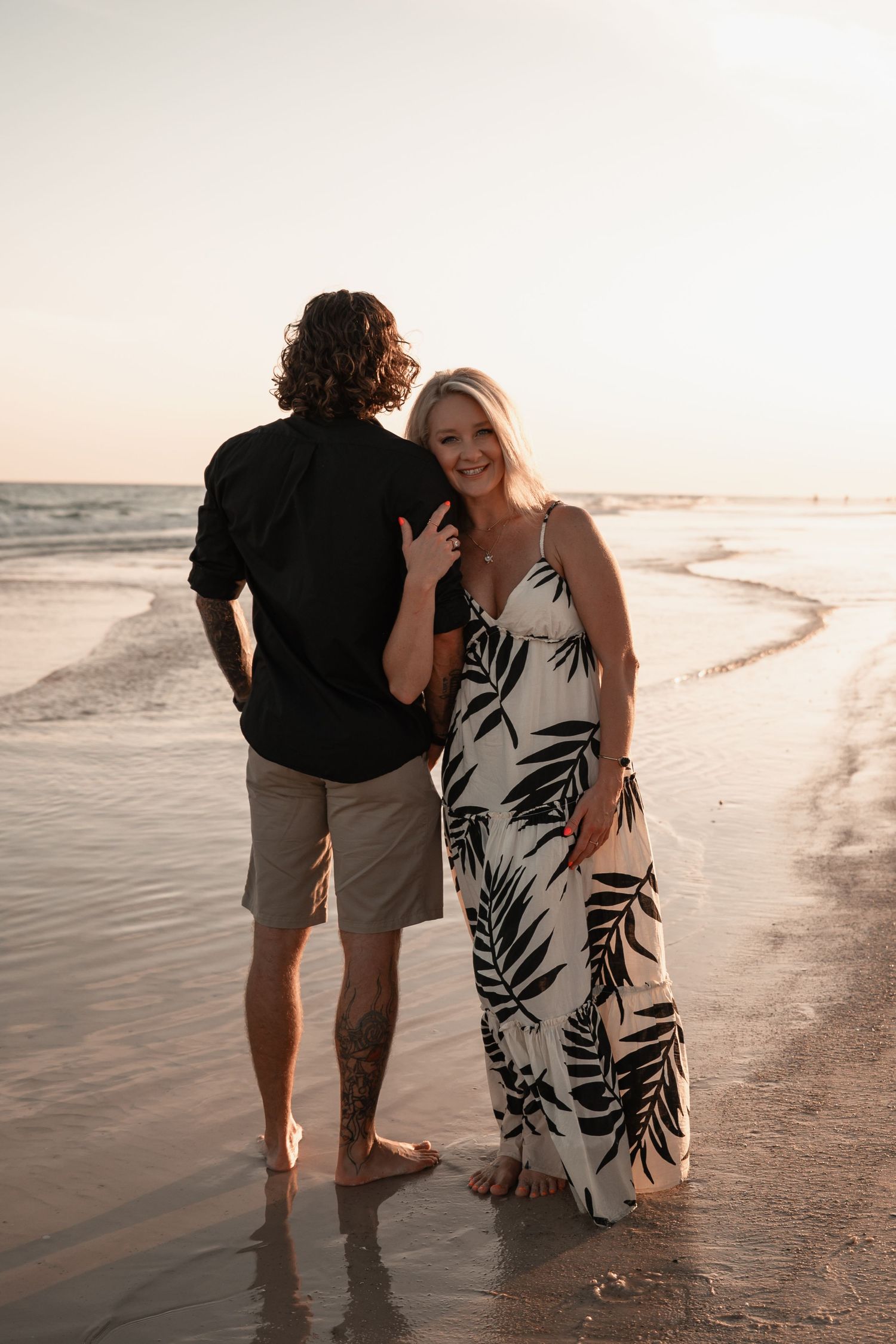 Couple posing at sunset on the beach, woman in floral maxi dress smiling at camera while man faces the ocean.