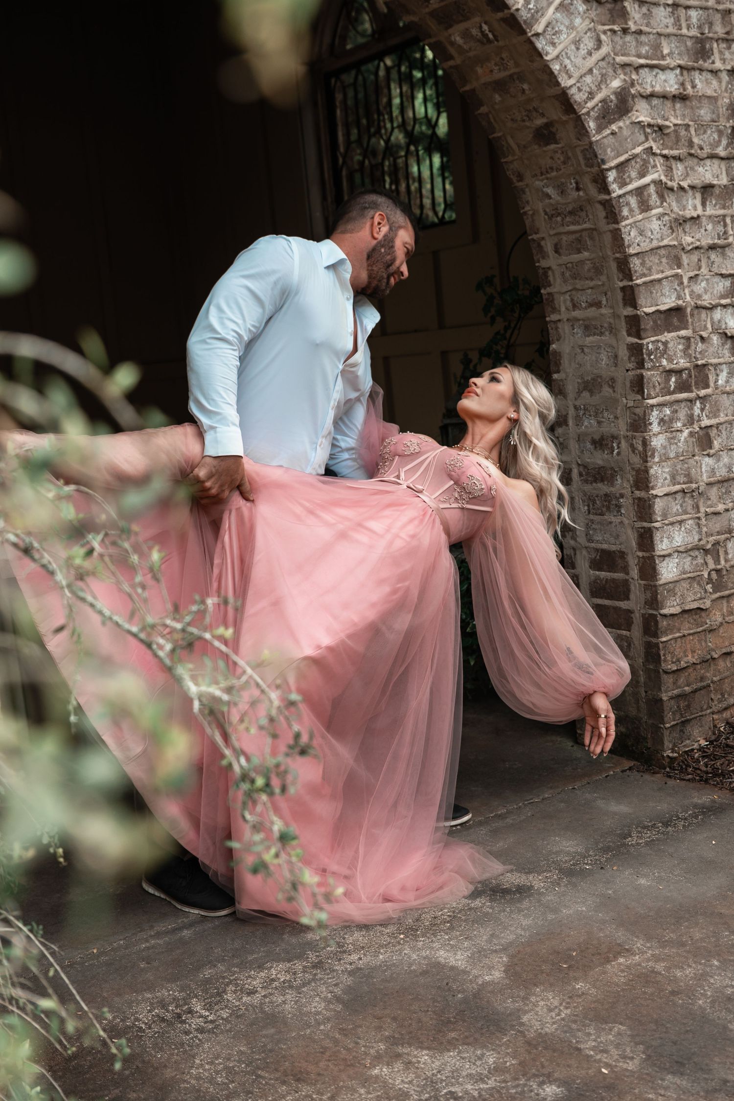 Man lifts woman in flowing pink tulle gown under a rustic brick archway in a romantic outdoor setting.