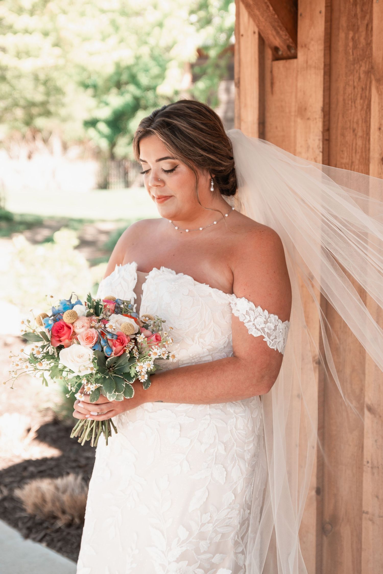 Elegant bride in lace off-shoulder wedding gown holding bouquet while leaning against rustic wooden wall.