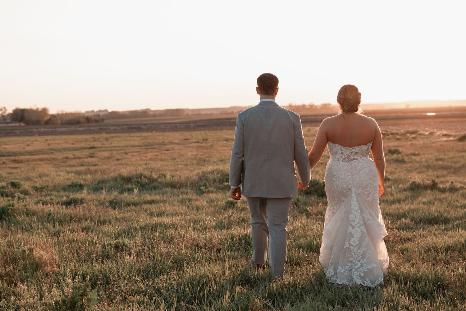 Bride and groom hold hands walking through golden field at sunset in elegant wedding attire.