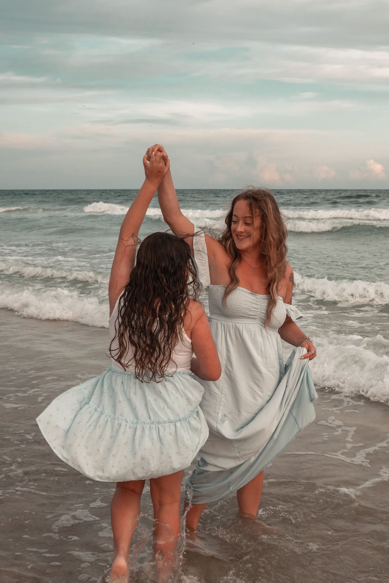 Two women in matching light blue dresses dance together on a beach with ocean waves behind them.