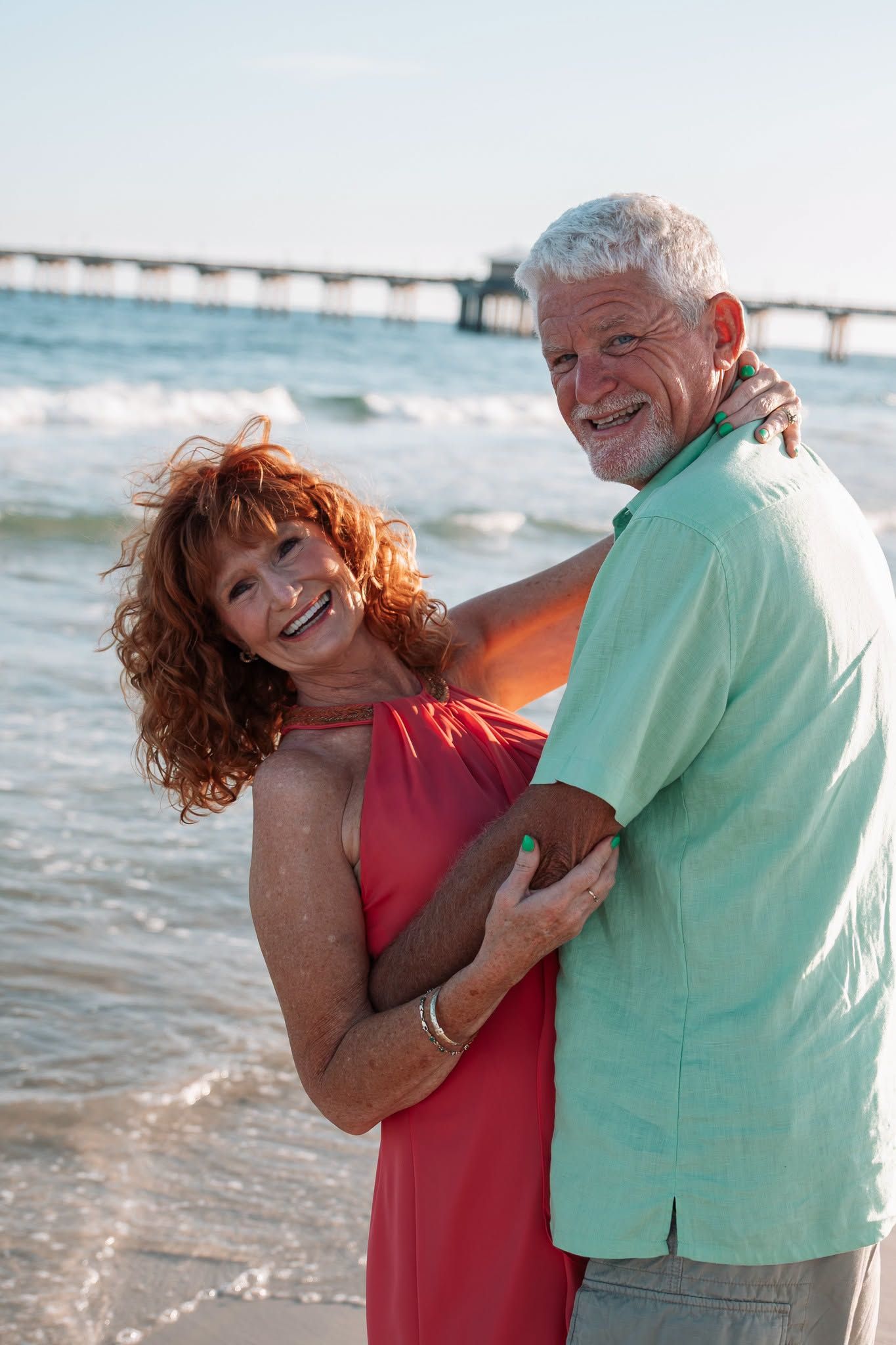 Happy senior couple embracing on a sunny beach near a pier, woman in red dress and man in mint green shirt.