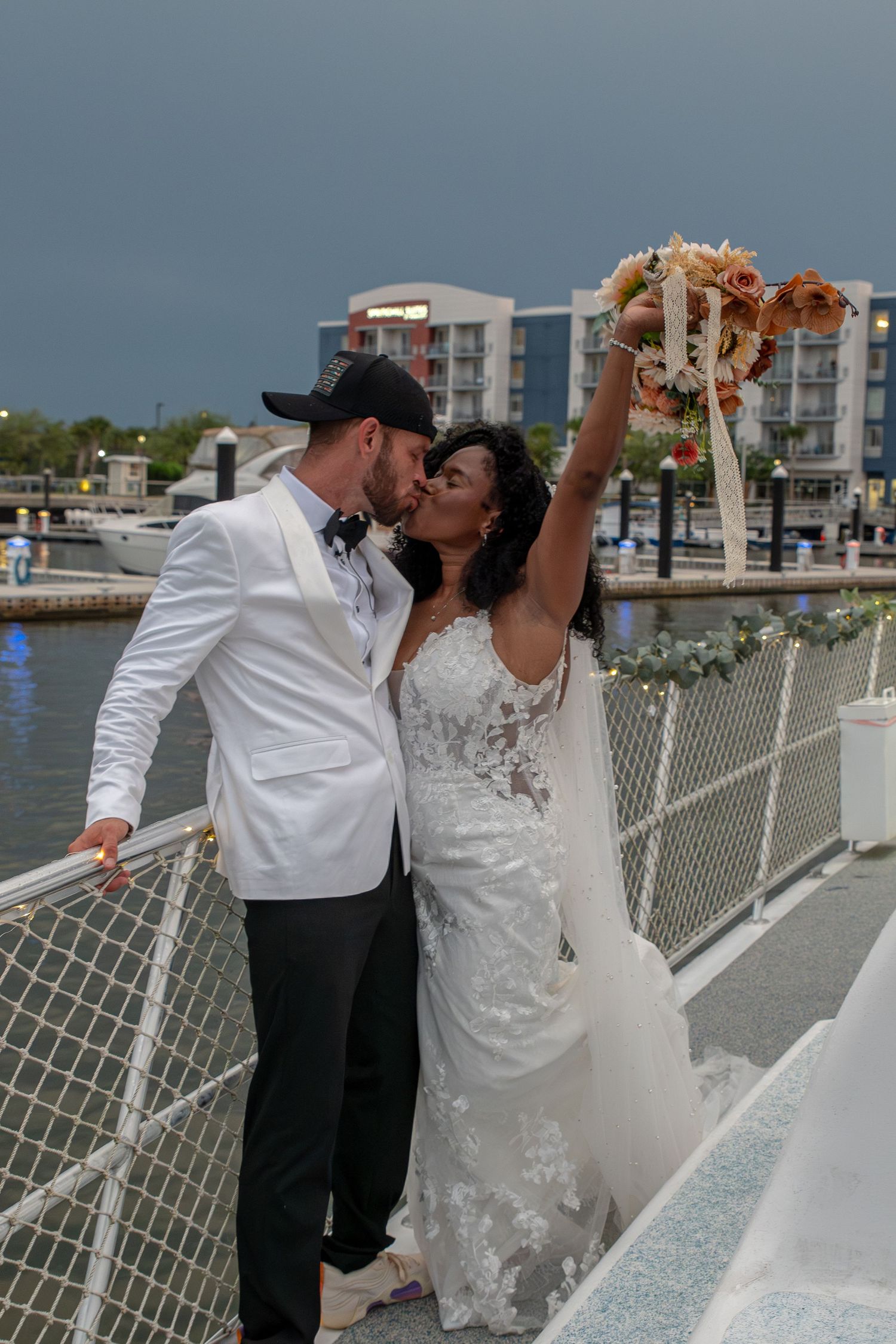 Bride and groom share a kiss on a waterfront dock, bride raising bouquet, groom in white tuxedo jacket and black cap.