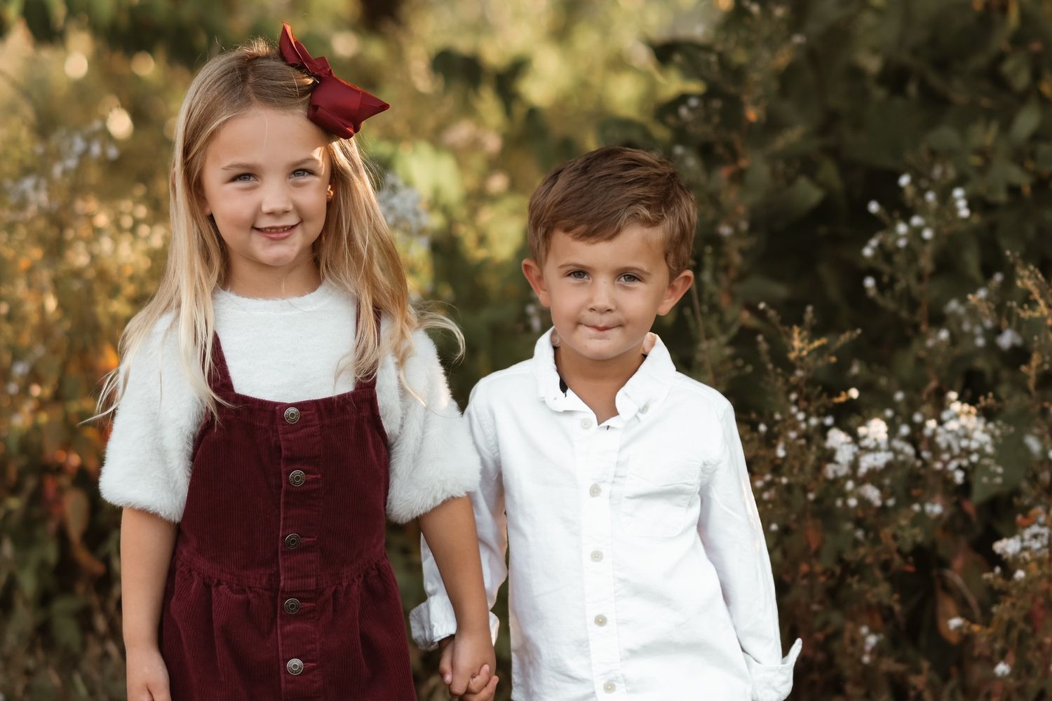 Young girl and boy holding hands outdoors, wearing coordinating burgundy and white outfits.