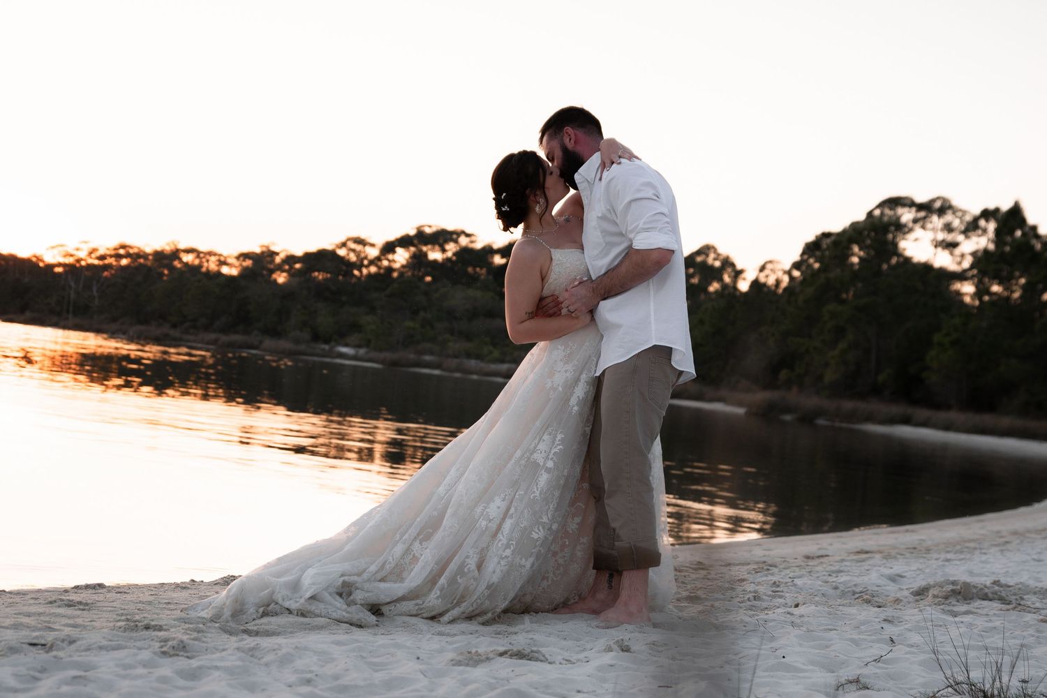 Bride and groom kissing on sandy beach at golden sunset by calm river.
