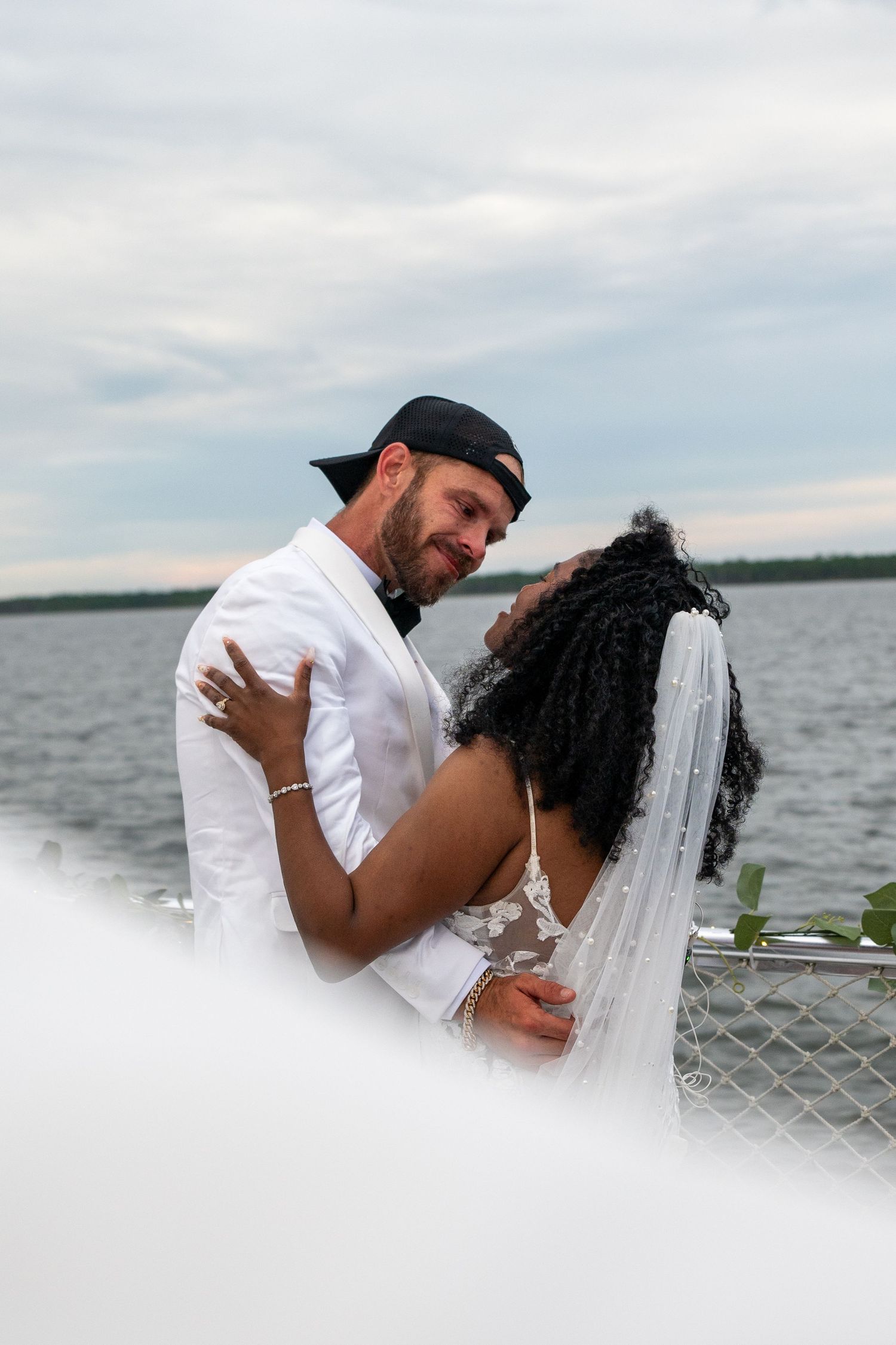 Interracial couple embracing on a boat deck in wedding attire, bride in lace gown with veil, groom in white suit with black cap.