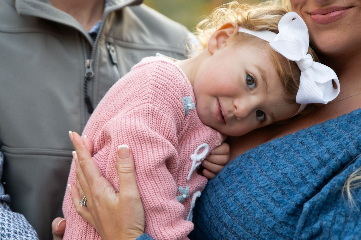 Adorable baby girl in pink sweater with white bow rests on mothers shoulder outdoors.