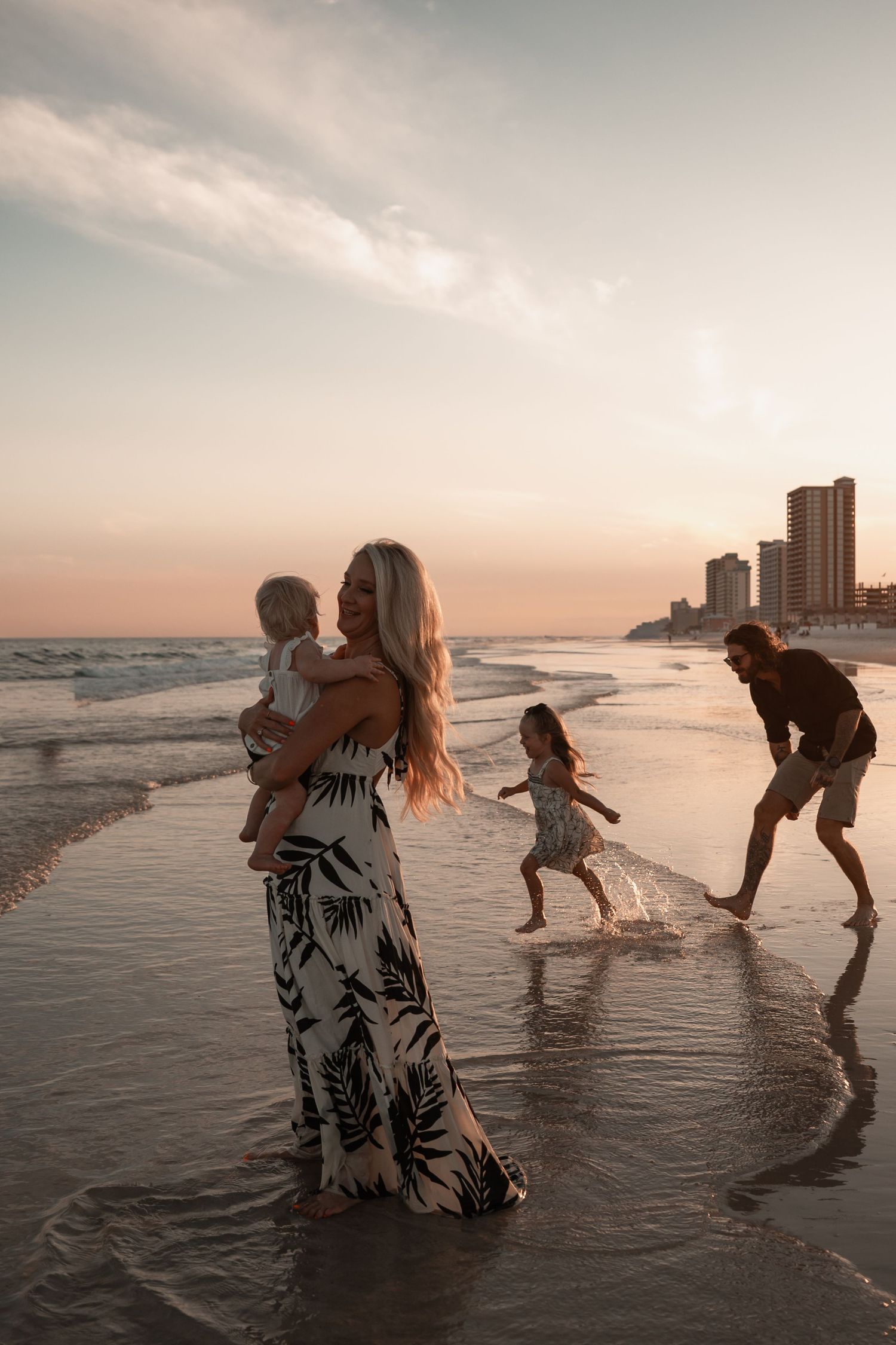 Family of four enjoying sunset on a beach with mother holding baby and father chasing daughter.