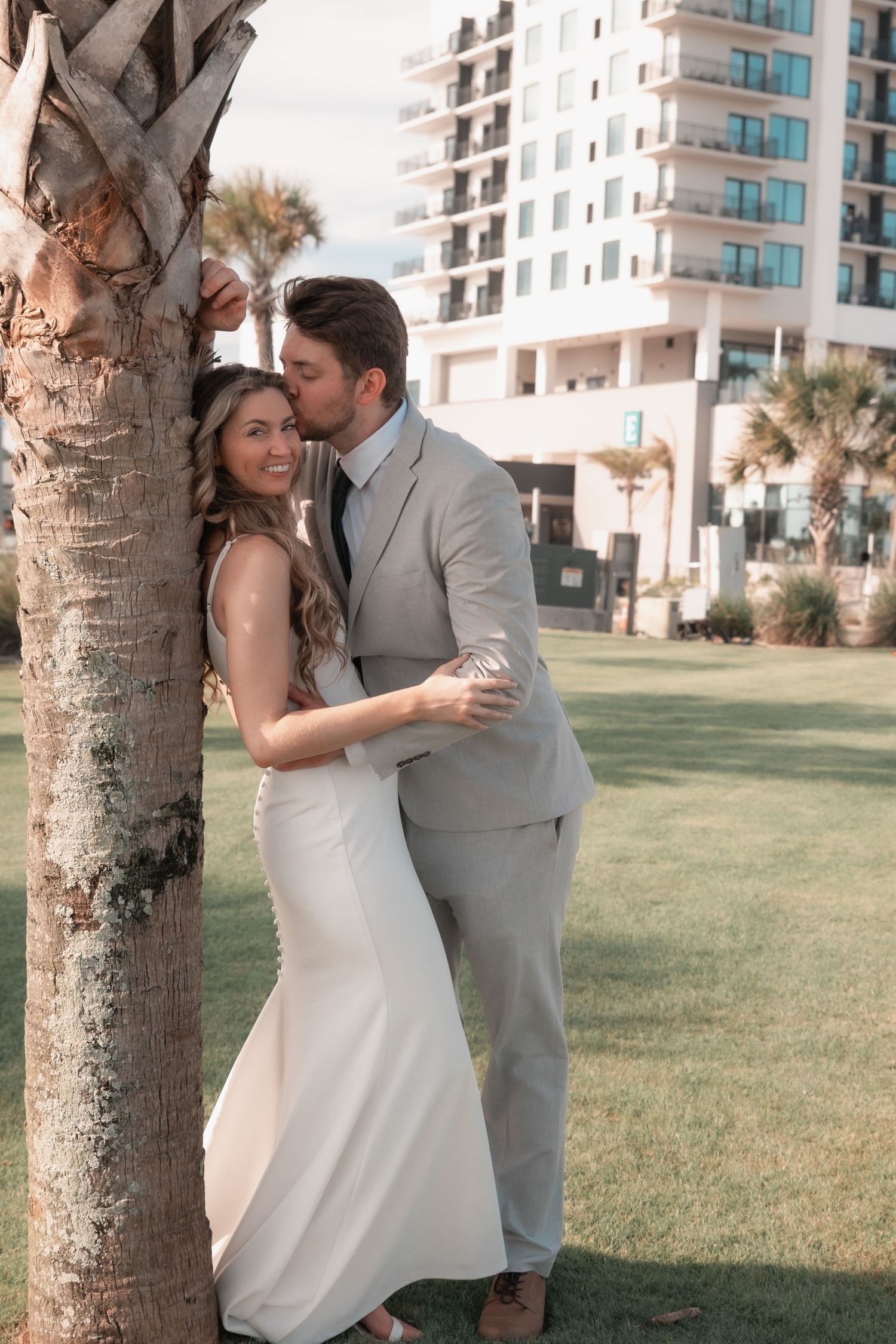 Groom kisses bride beside a palm tree with coastal hotel in background.