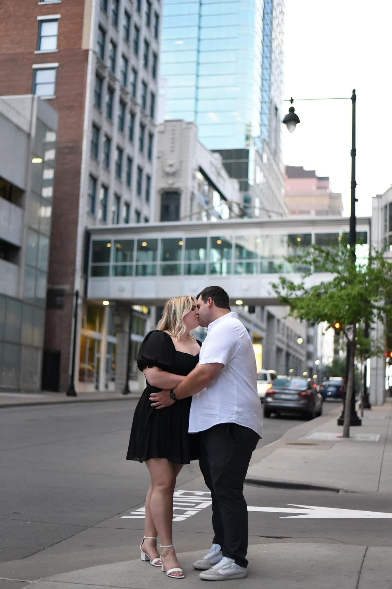 Couple sharing a passionate kiss on a city street corner surrounded by tall urban buildings at dusk.
