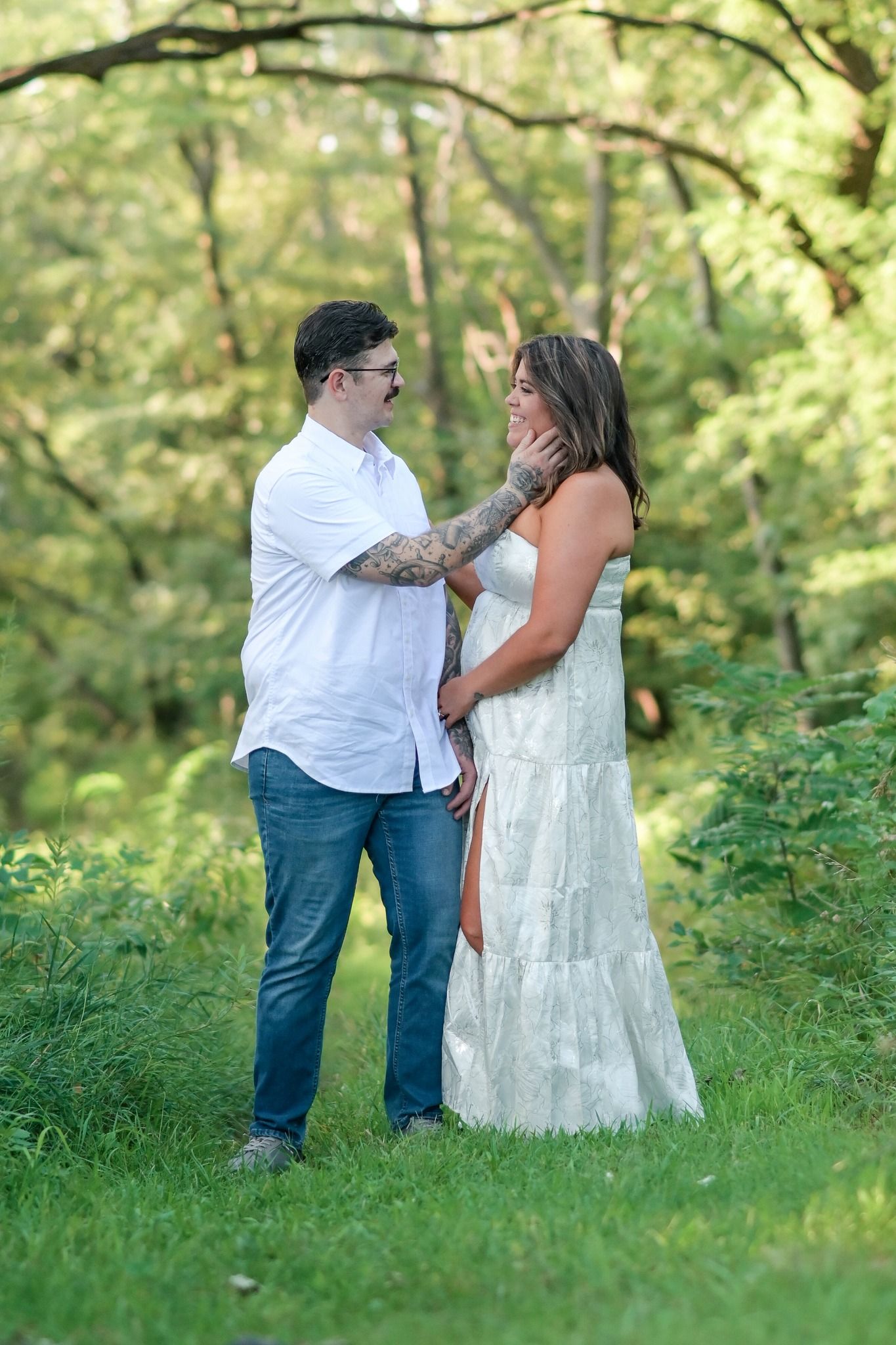 Couple standing in green forest, man with tattoos tenderly cupping womans face in white lace dress.
