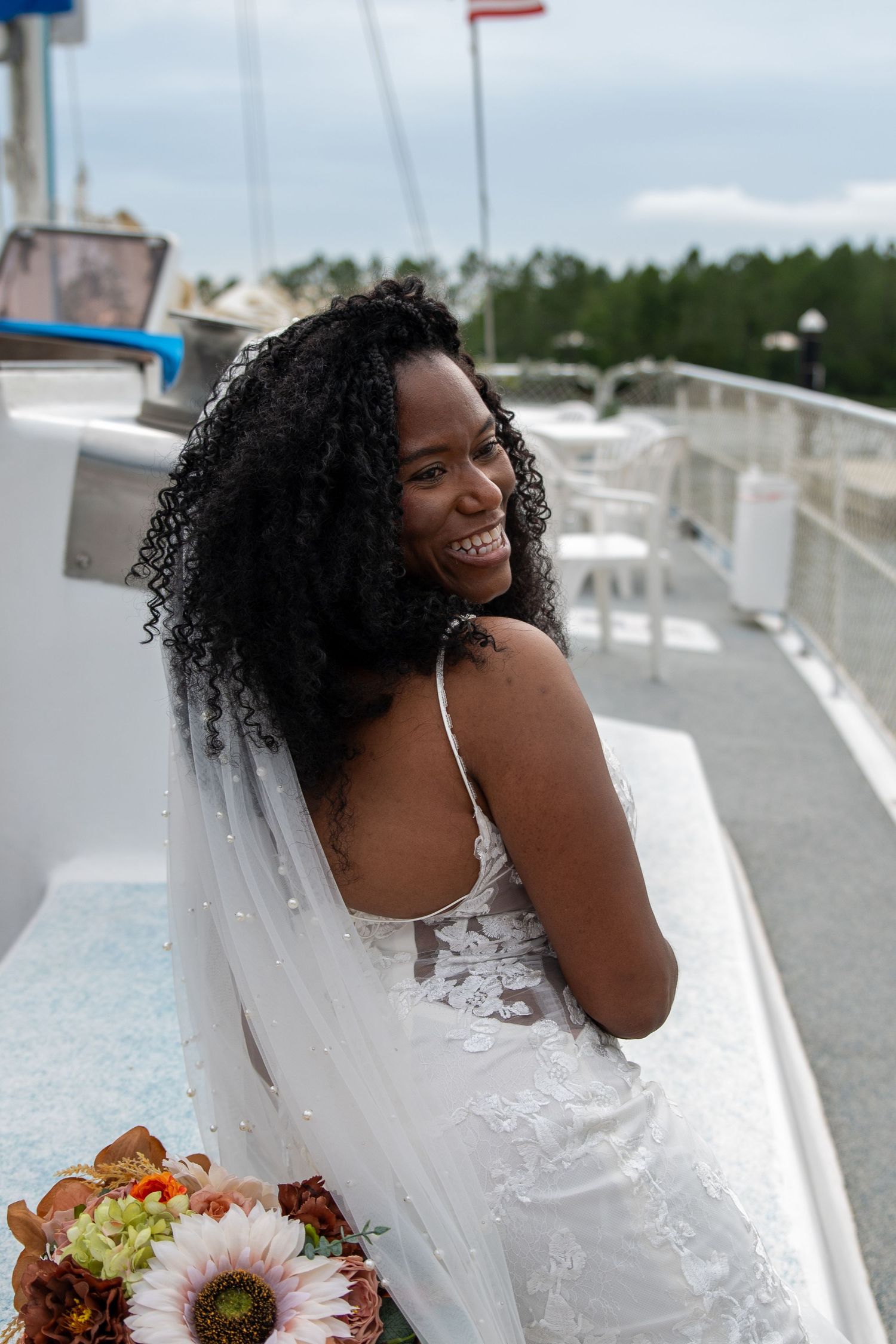 Smiling Black bride in white lace gown with veil holds colorful bouquet on boat deck, looking back over shoulder.