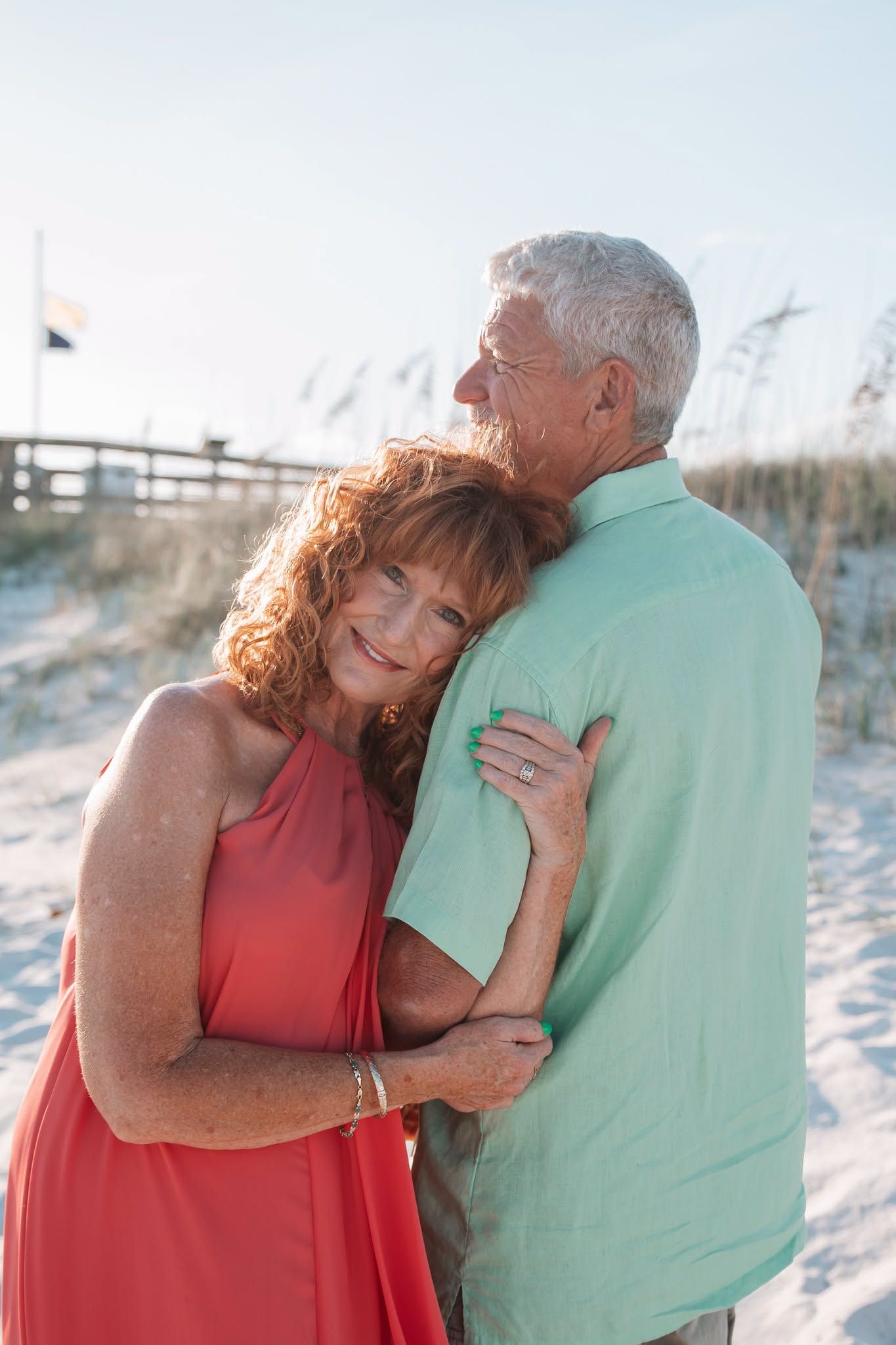 A happy older couple embraces on a sandy beach, the woman in a red dress smiling warmly as they share a romantic moment.