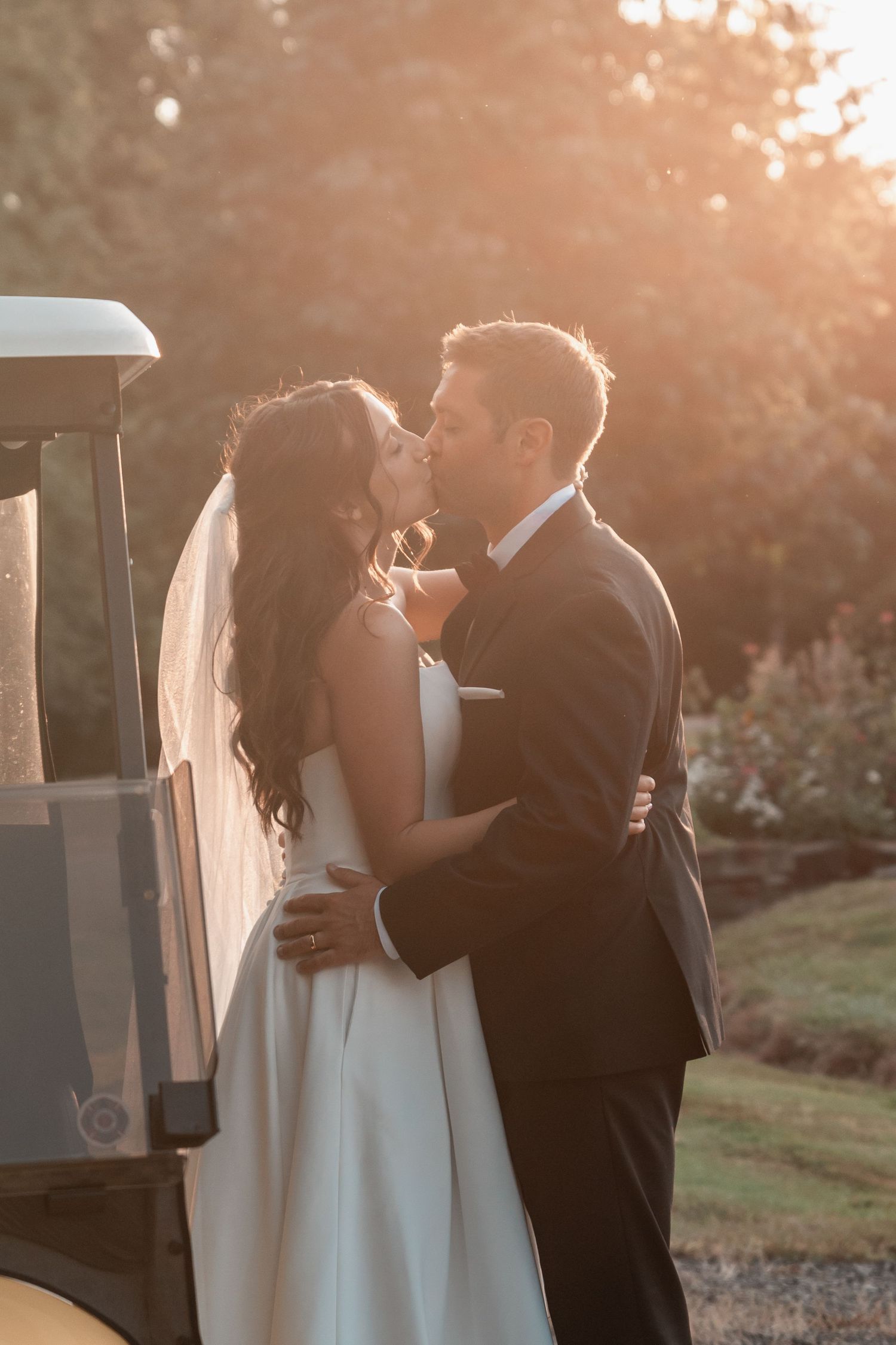Bride and groom share a golden hour kiss in wedding attire, with soft sunlight glowing behind them outdoors.