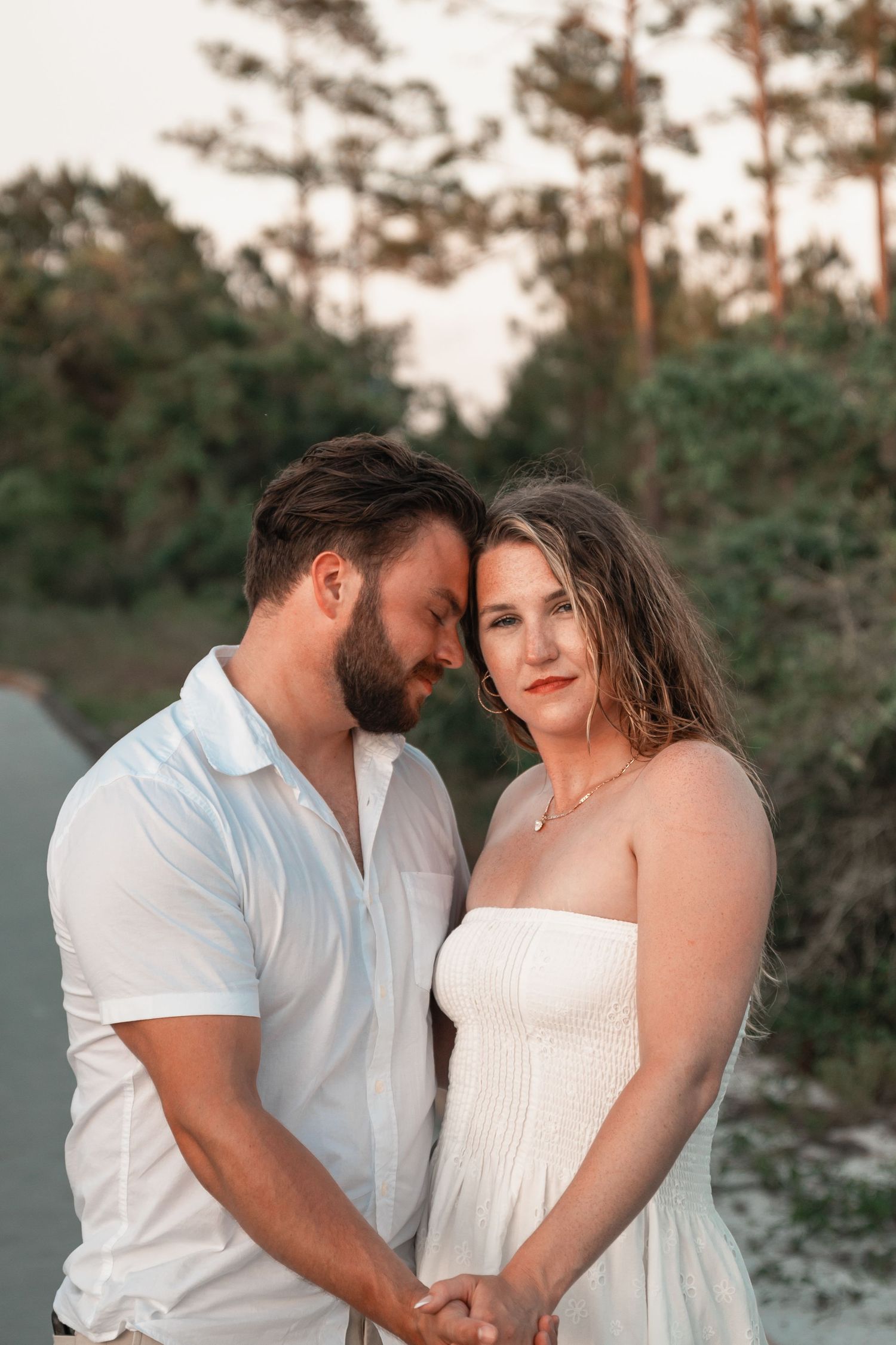 Engaged couple holding hands on sandy path at sunset, woman in white strapless dress leaning on mans shoulder.