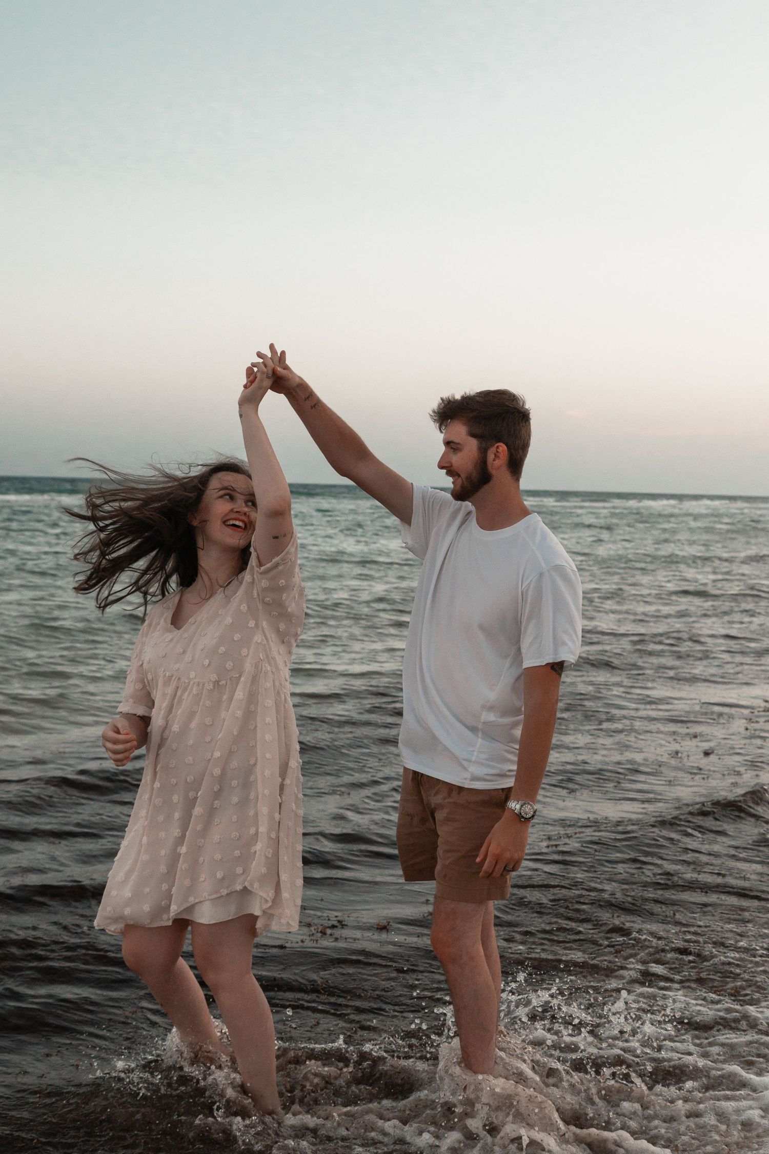 A joyful couple dancing in ocean waves at sunset, man in white tee spinning woman in flowy dress on beach.