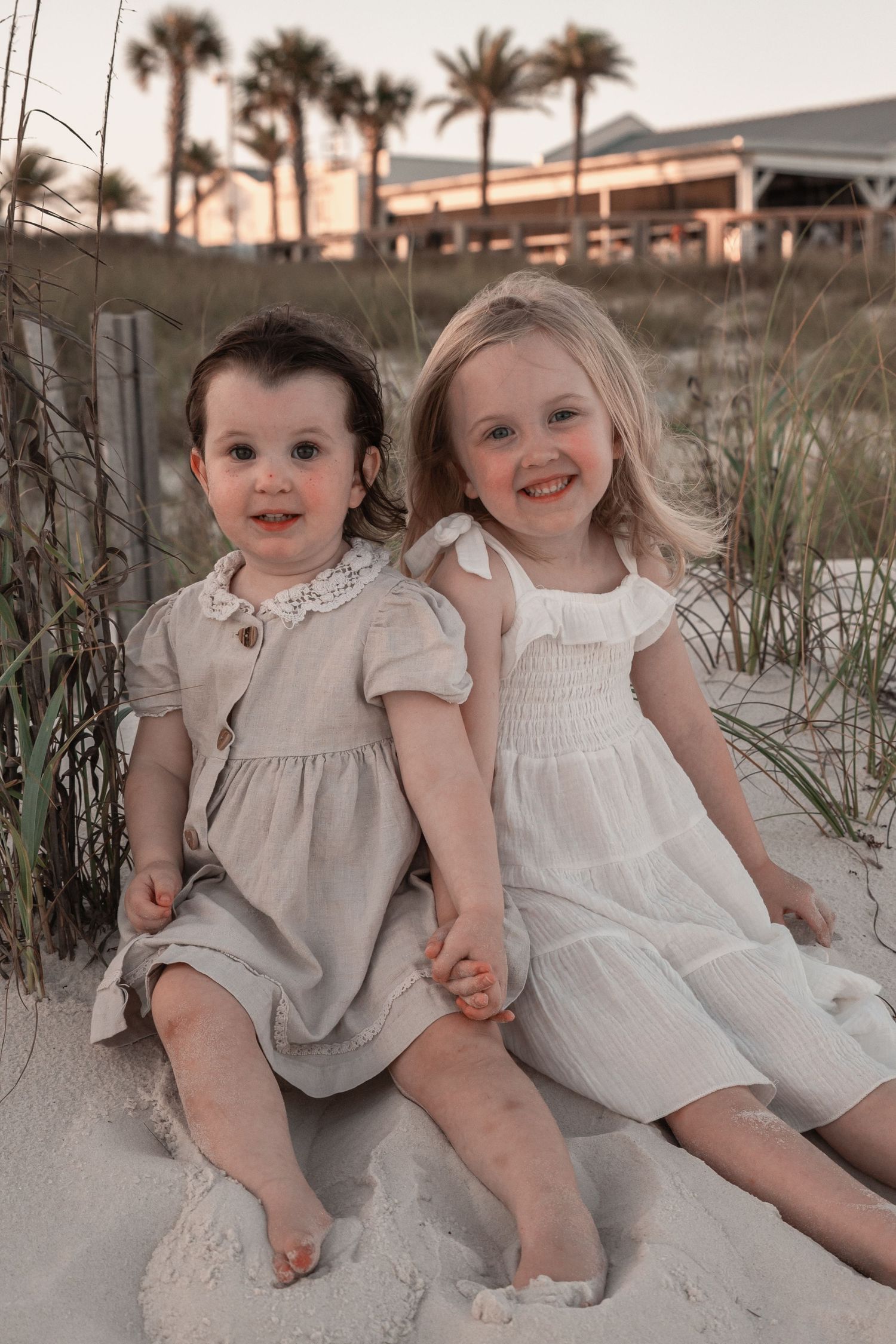 Two young girls in matching linen dresses sit together on sandy beach dunes at sunset, smiling and holding hands.