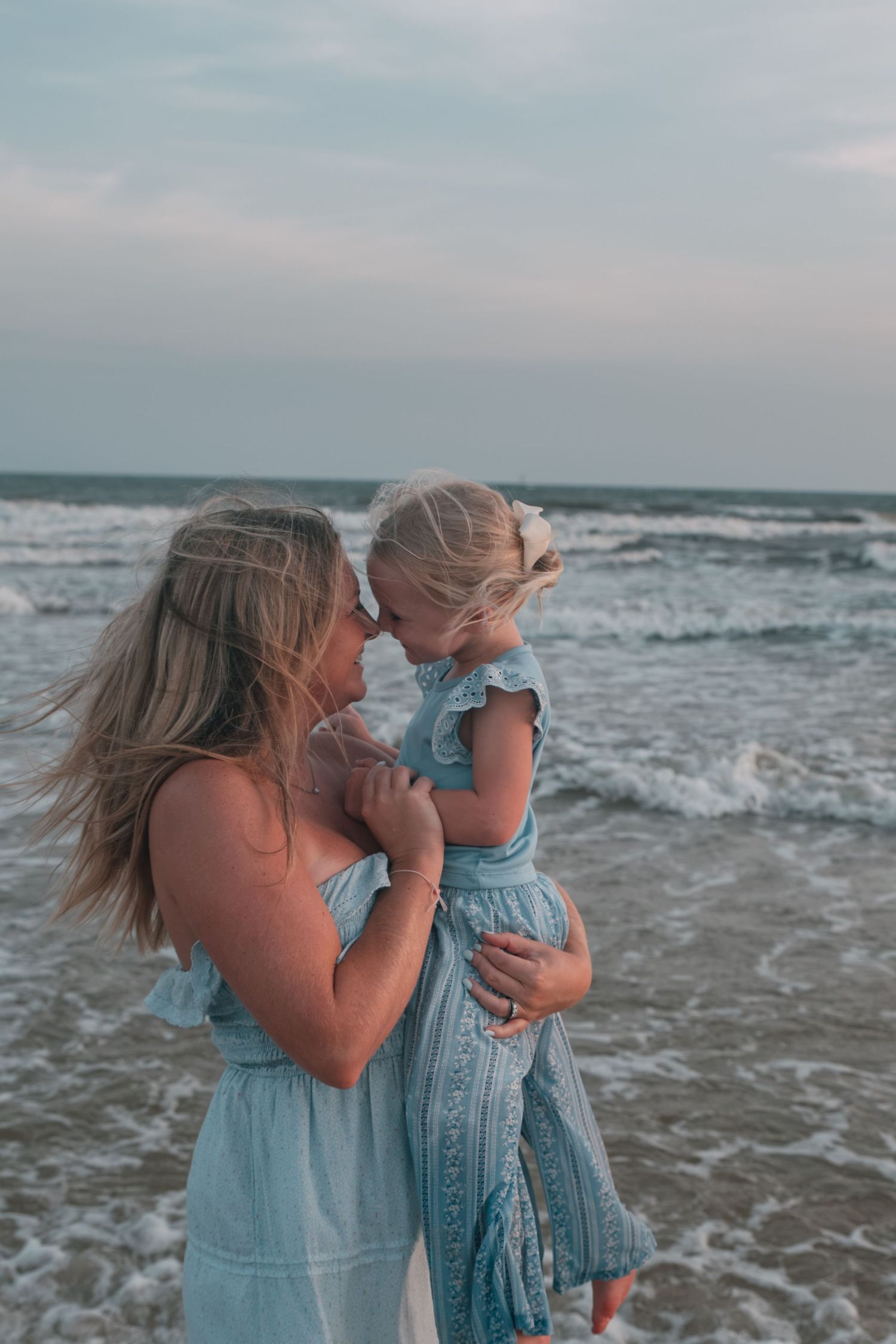 A mother and young daughter in matching light blue dresses share a tender moment on a beach with ocean waves behind them.
