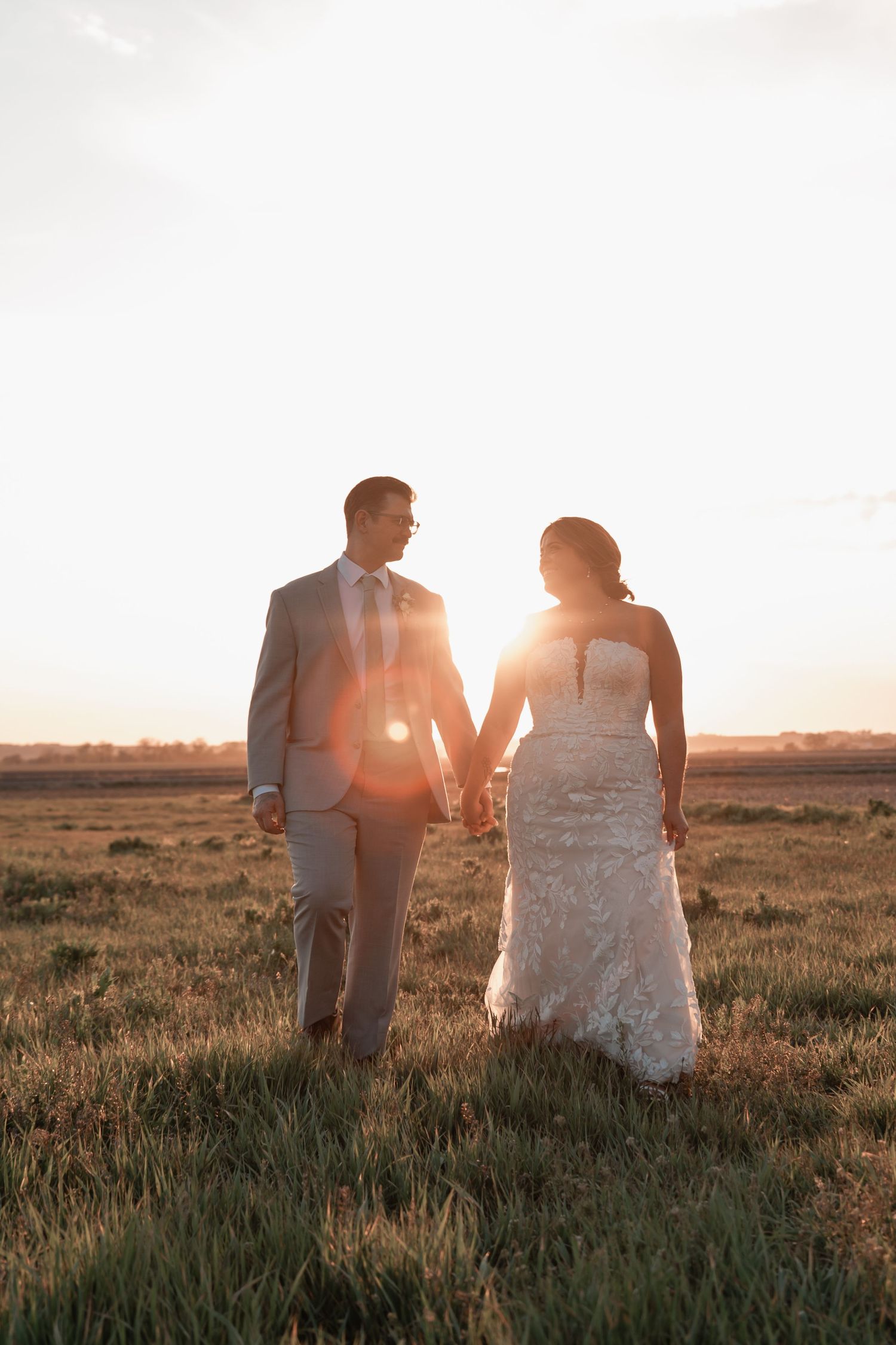 Recently married couple walking hand in hand through a grassy field under soft sunset light