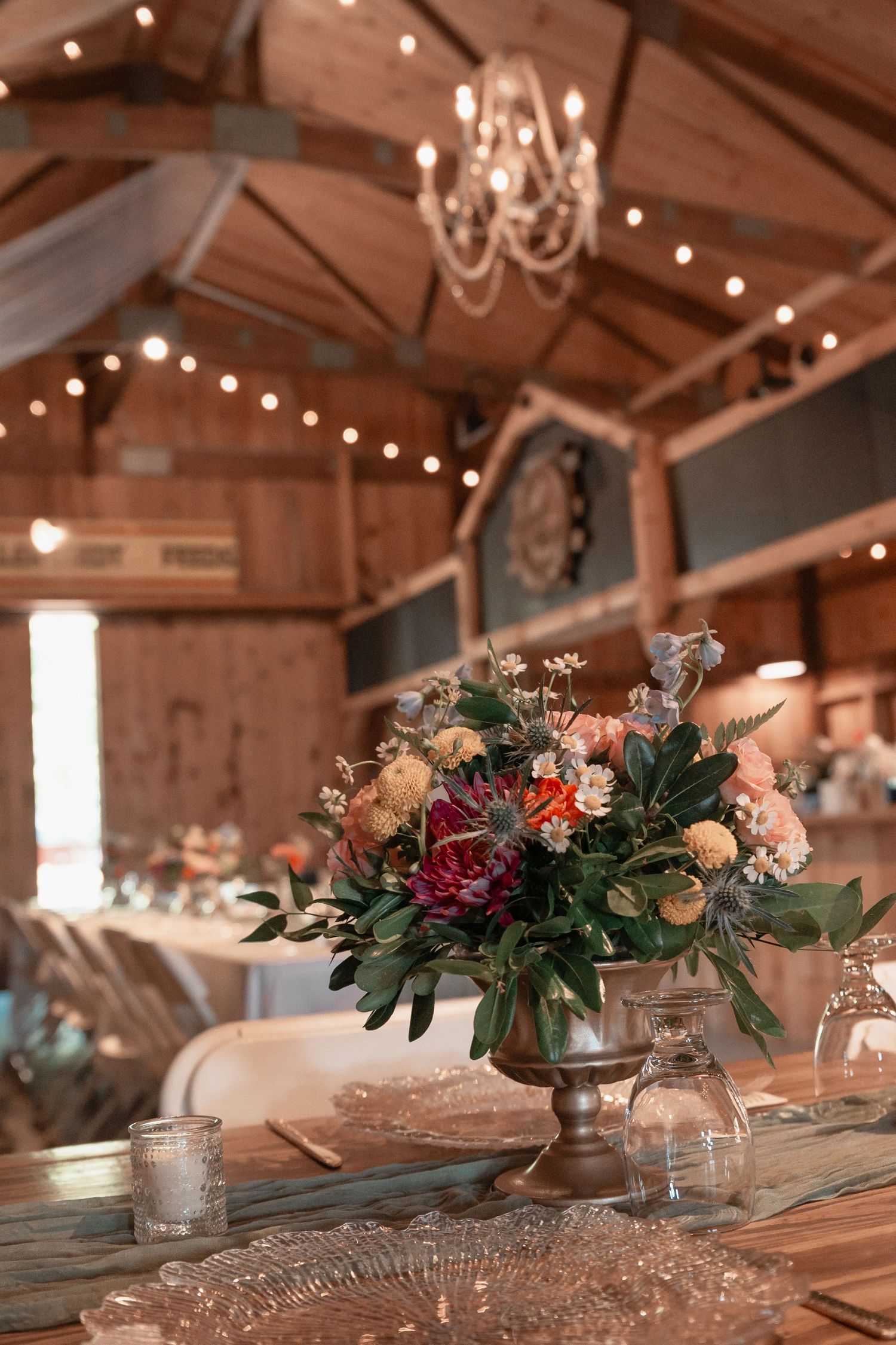 Rustic barn wedding reception with colorful floral centerpiece, chandelier, and warm string lights overhead.