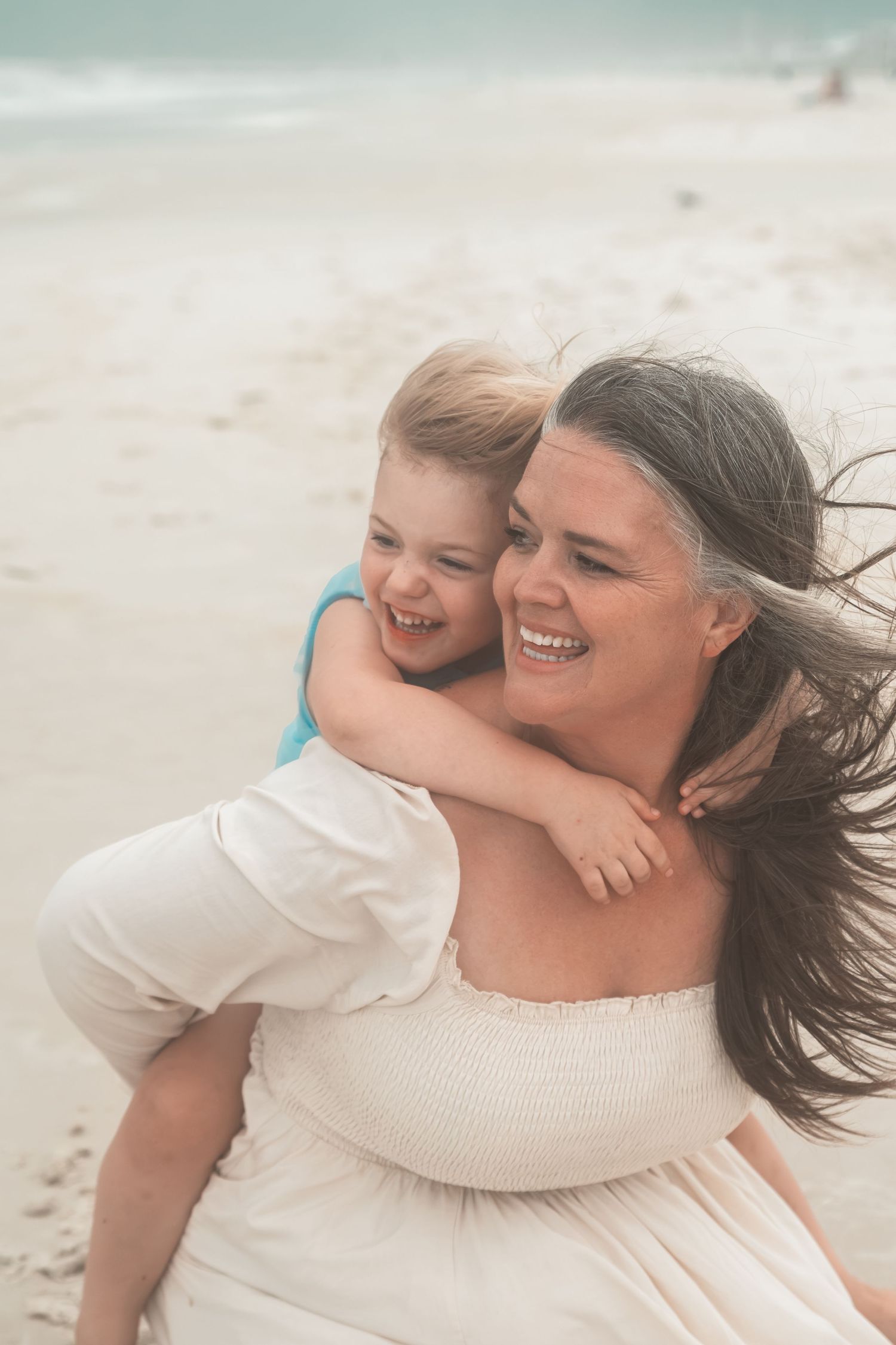 A joyful woman in a white dress gives a laughing child a piggyback ride on a sandy beach.