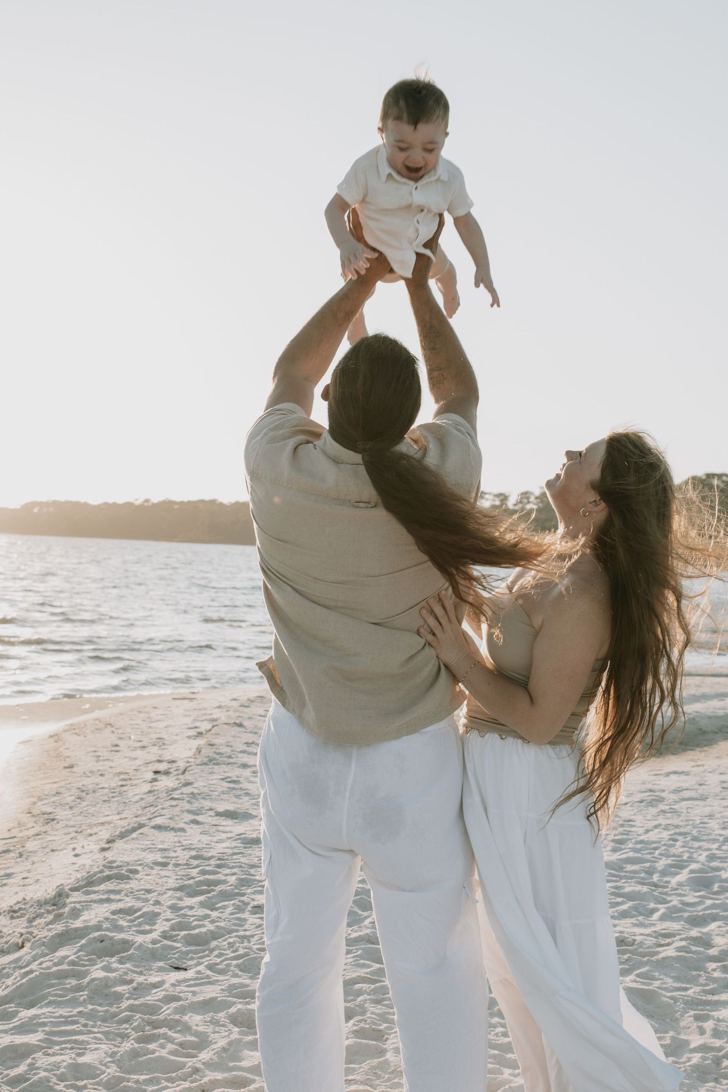 Parents lifting smiling baby in the air on a sunlit beach.