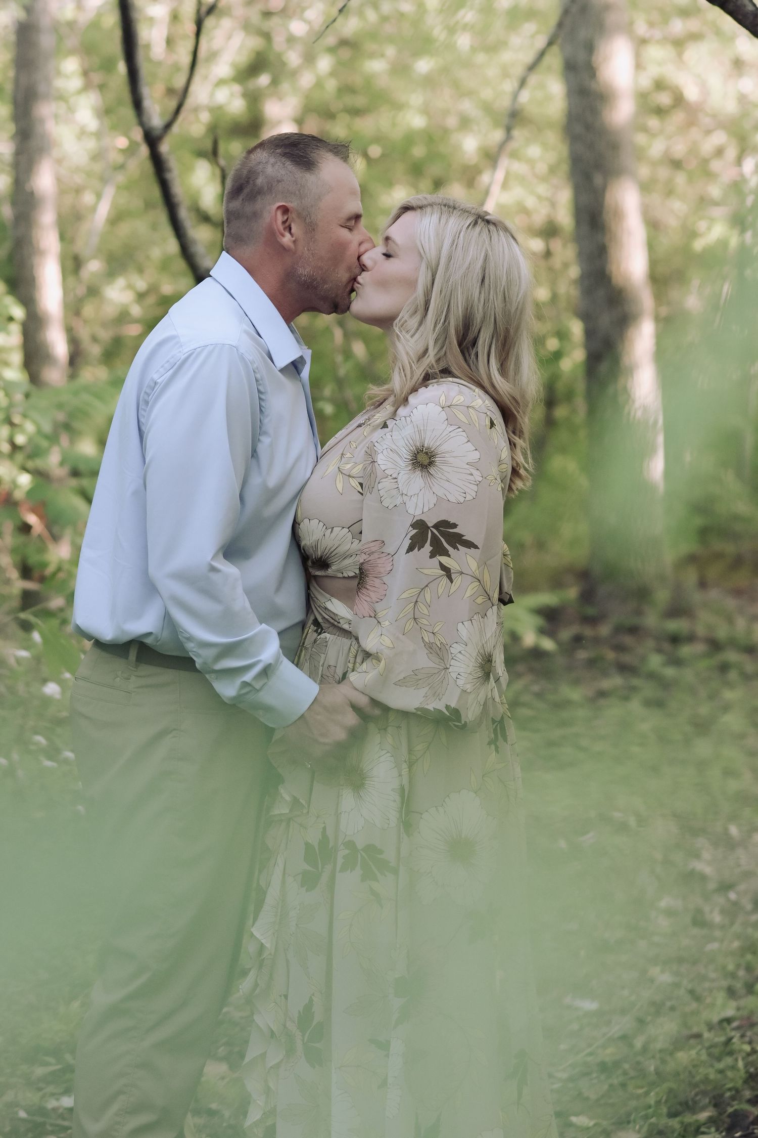 Engaged couple sharing a kiss among green trees, woman in elegant floral maxi dress holding hands with partner.