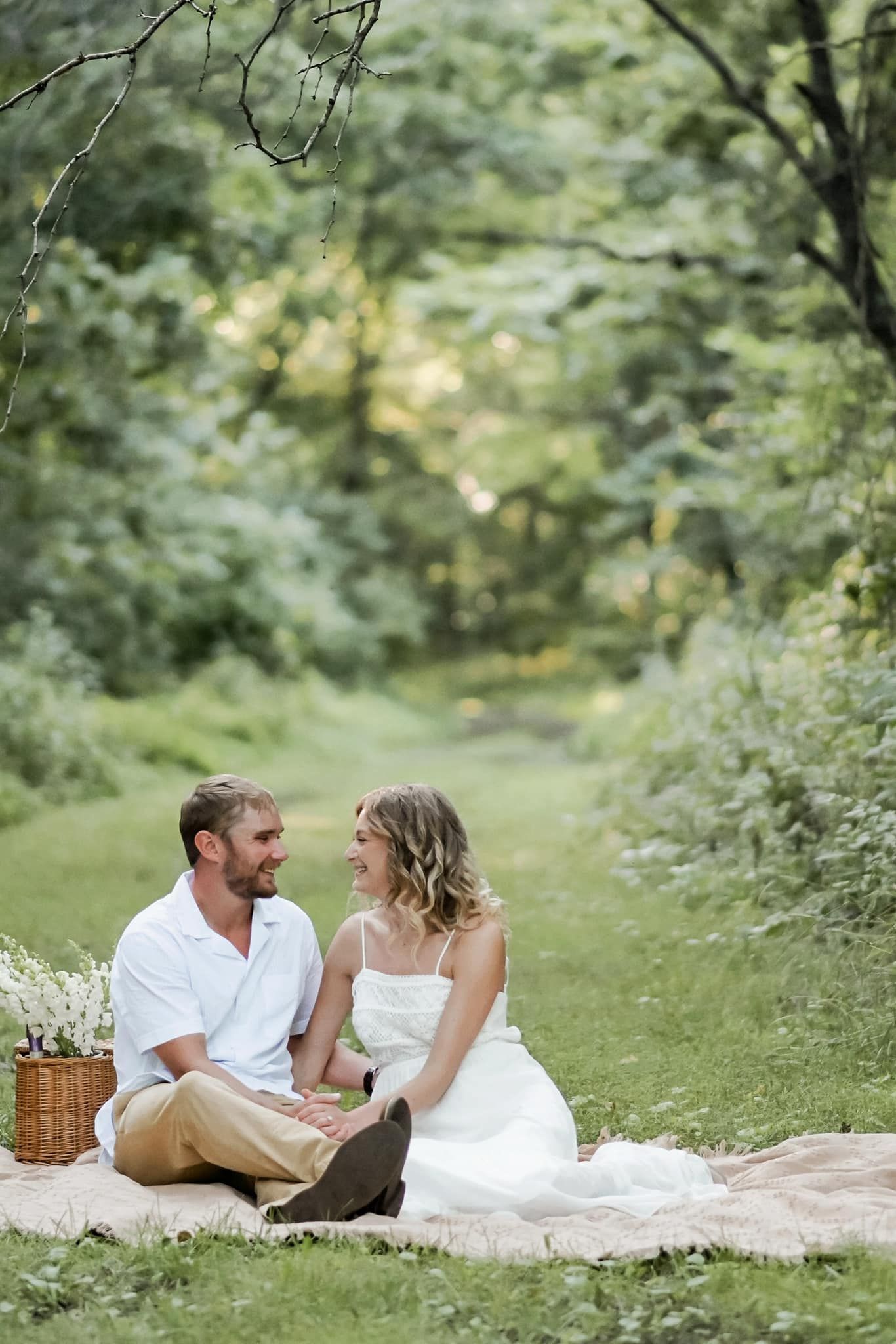 Couple sitting on a blanket in a lush green forest, smiling at each other during a romantic picnic.