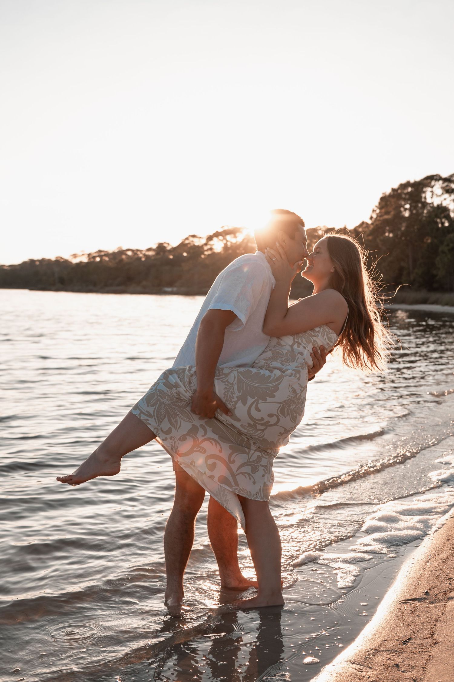 Man lifting woman in floral dress at sunset beach, sharing romantic kiss.