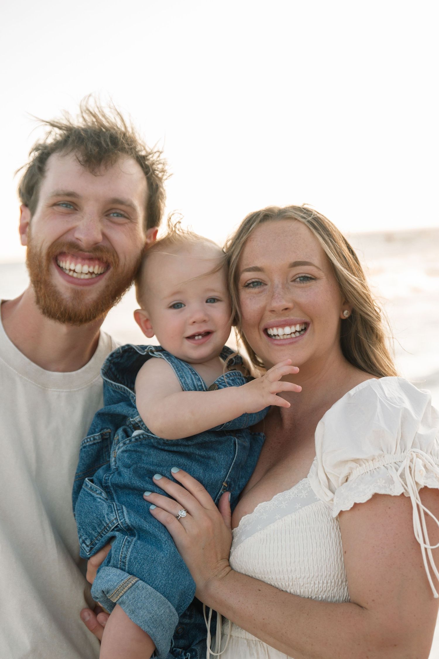 Happy family of three smiling at beach, parents holding baby in denim overalls, golden hour sunset backdrop.