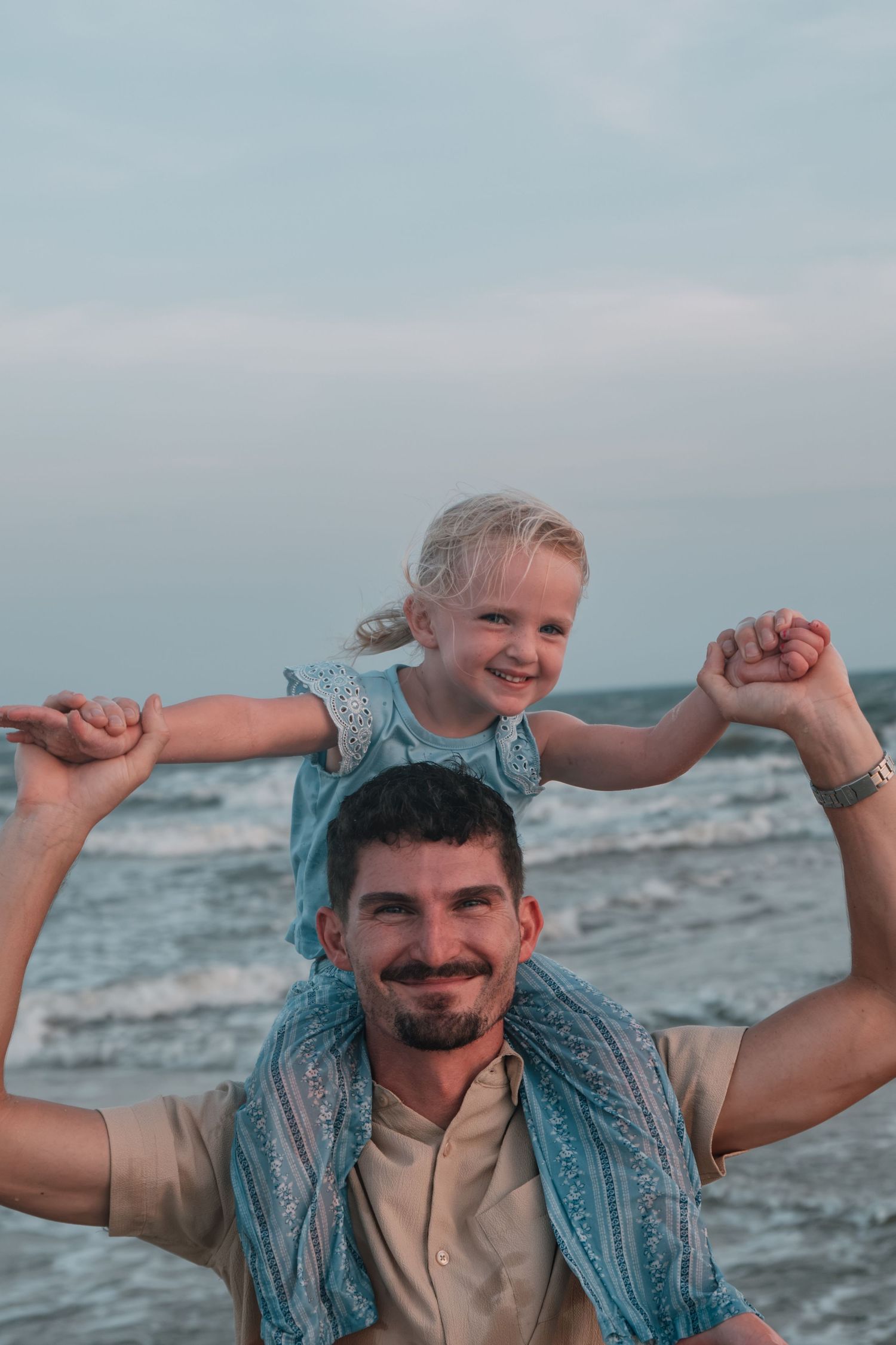 A smiling man carries a happy blonde girl on his shoulders at the beach, both enjoying a playful moment by the ocean.